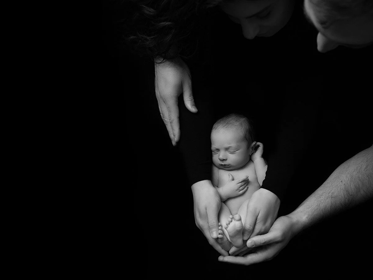 Black and white photo of a newborn baby being held gently by two adults, one supporting the baby's feet and the other cradling the baby's head and body, against a dark background.