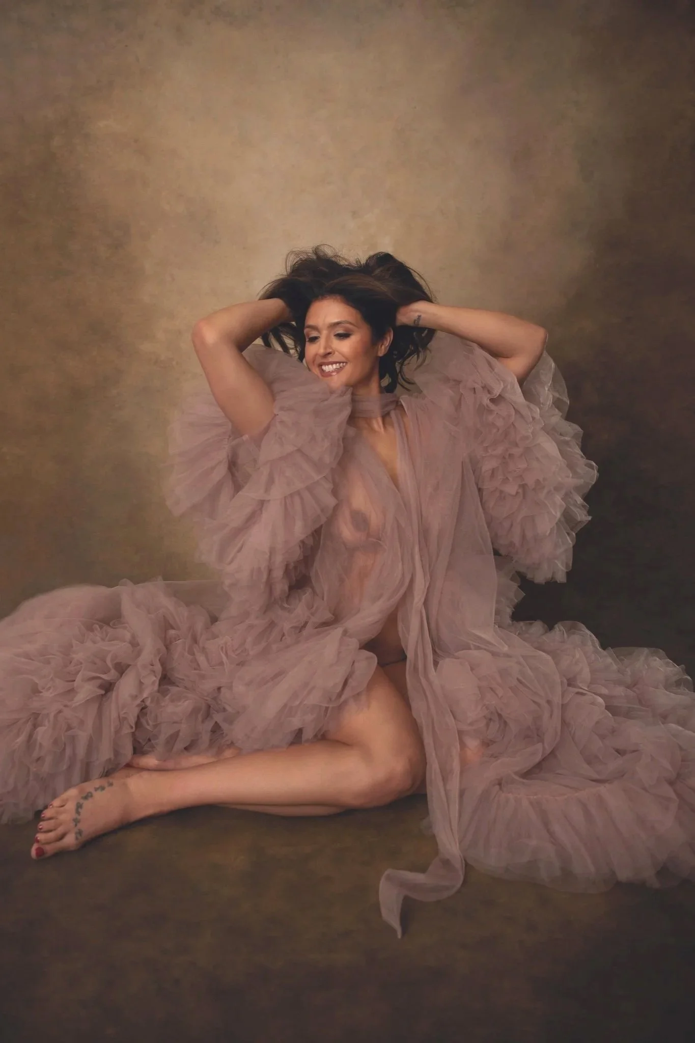 A woman with dark hair, smiling and lying on her back on a dark surface, wearing a pink, ruffled, flowing dress, with her arms raised and hands in her hair, with one leg bent and her foot visible, displaying a tattoo on her ankle.