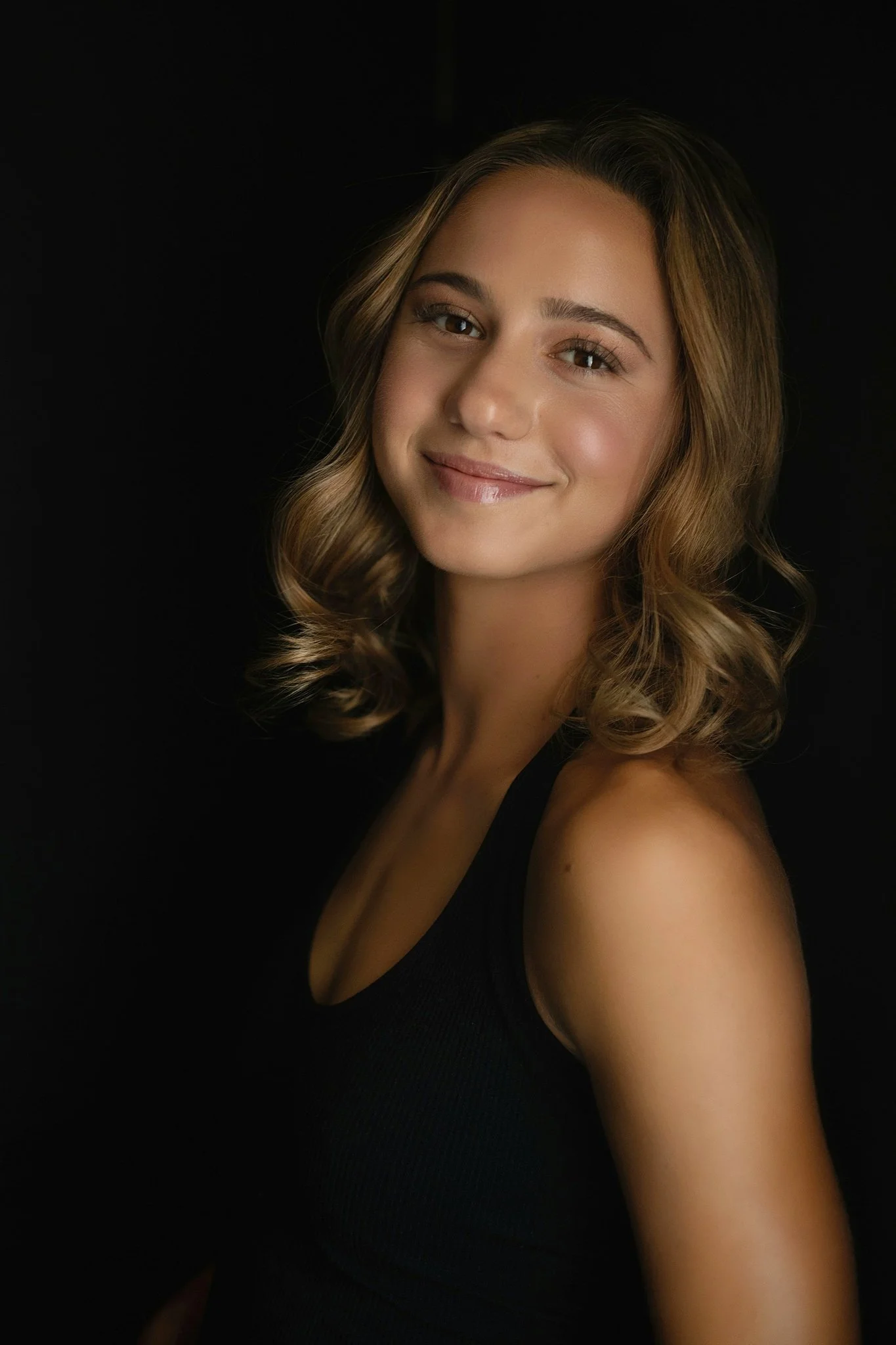A young woman with light brown hair styled in loose curls, smiling, wearing a black sleeveless top, with a dark background.