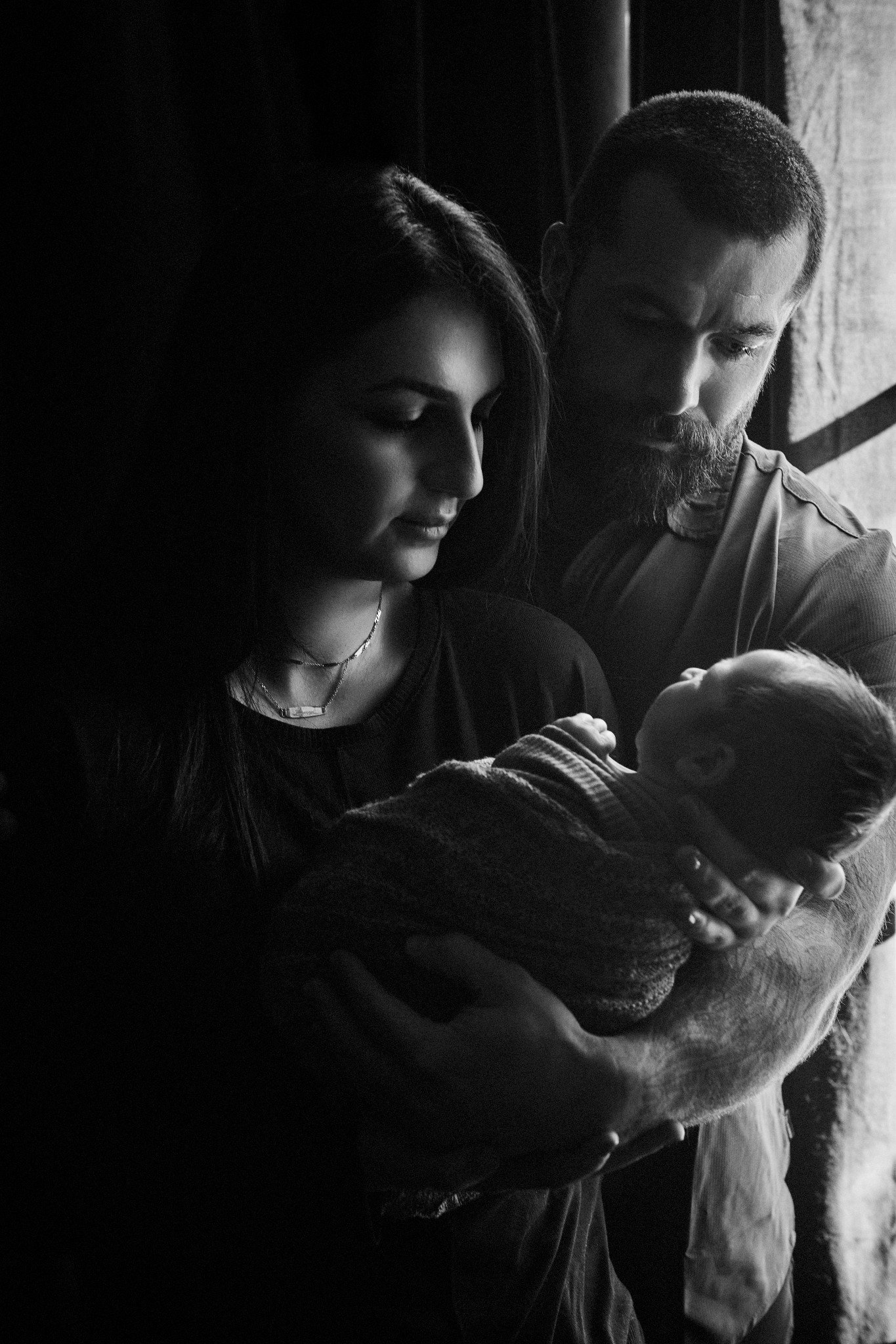 A black and white photo of a woman and a man looking down at a baby, with the woman on the left and the man on the right.