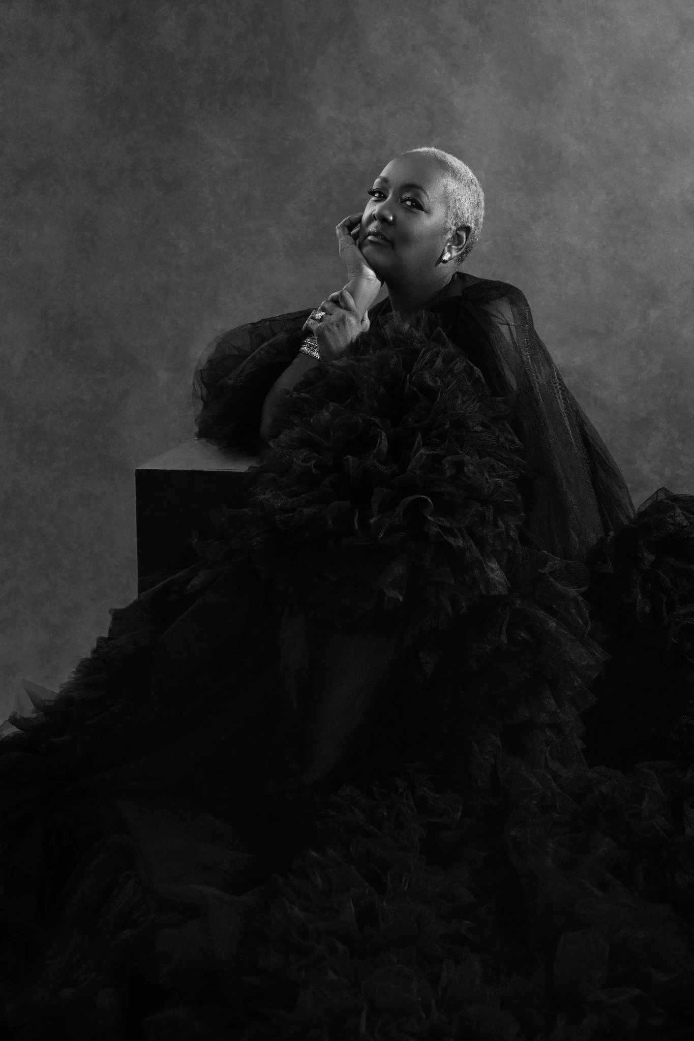 A black-and-white portrait of a woman with short, light-colored hair wearing a dark, ruffled dress. She is seated with her chin resting on her hand, looking confidently at the camera.