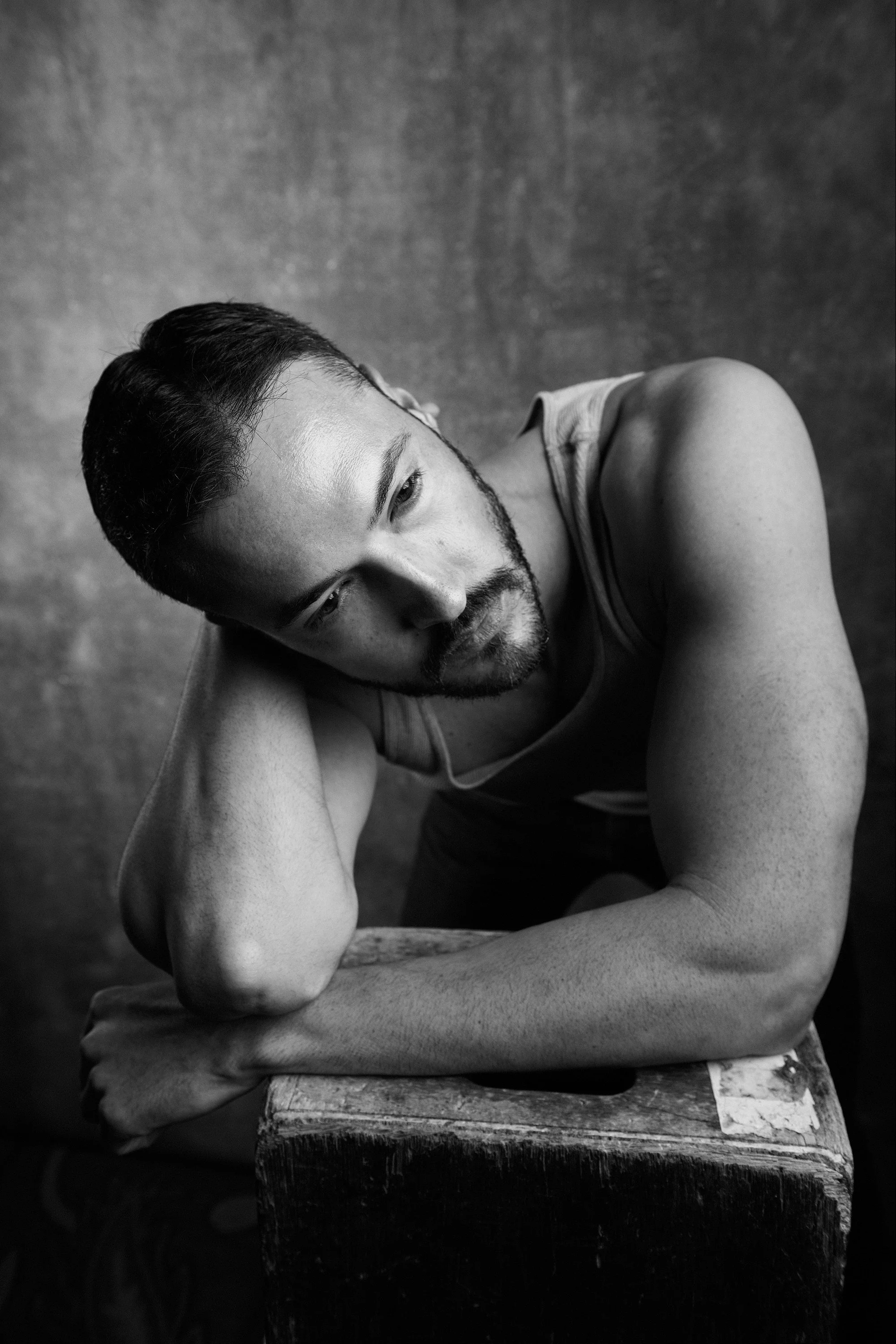 A black and white portrait of a man with short dark hair and a beard, resting his head on his arm, which is on a wooden surface, with a contemplative expression.