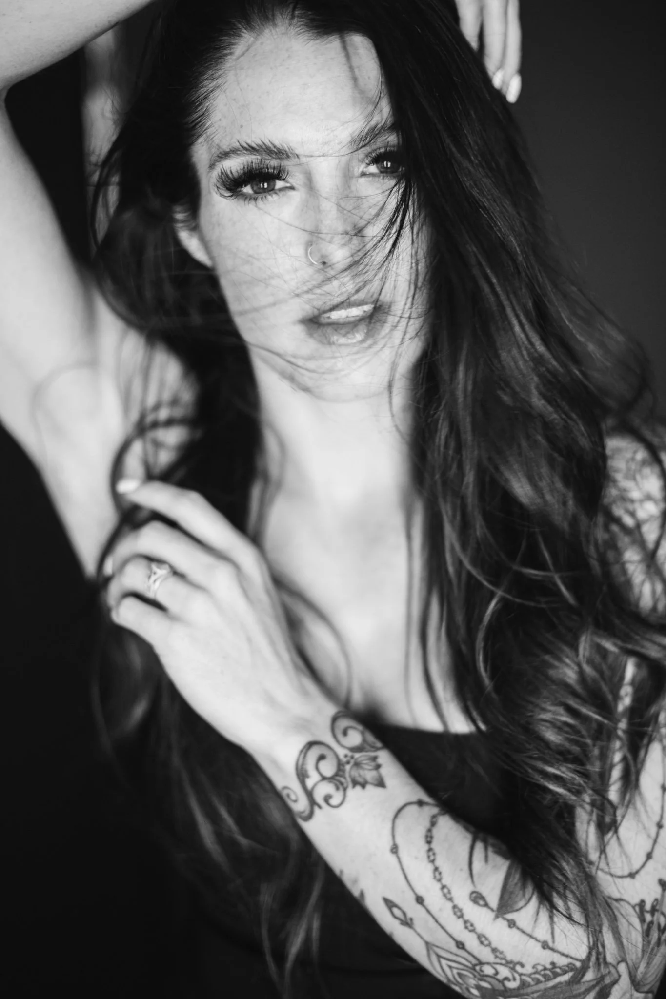 A black and white photograph of a woman with long, dark hair. She is looking directly at the camera with an intense expression, lips slightly parted, and hair blowing across her face. She has tattoos on her arm, a nose ring, and is touching her hair and shoulder.