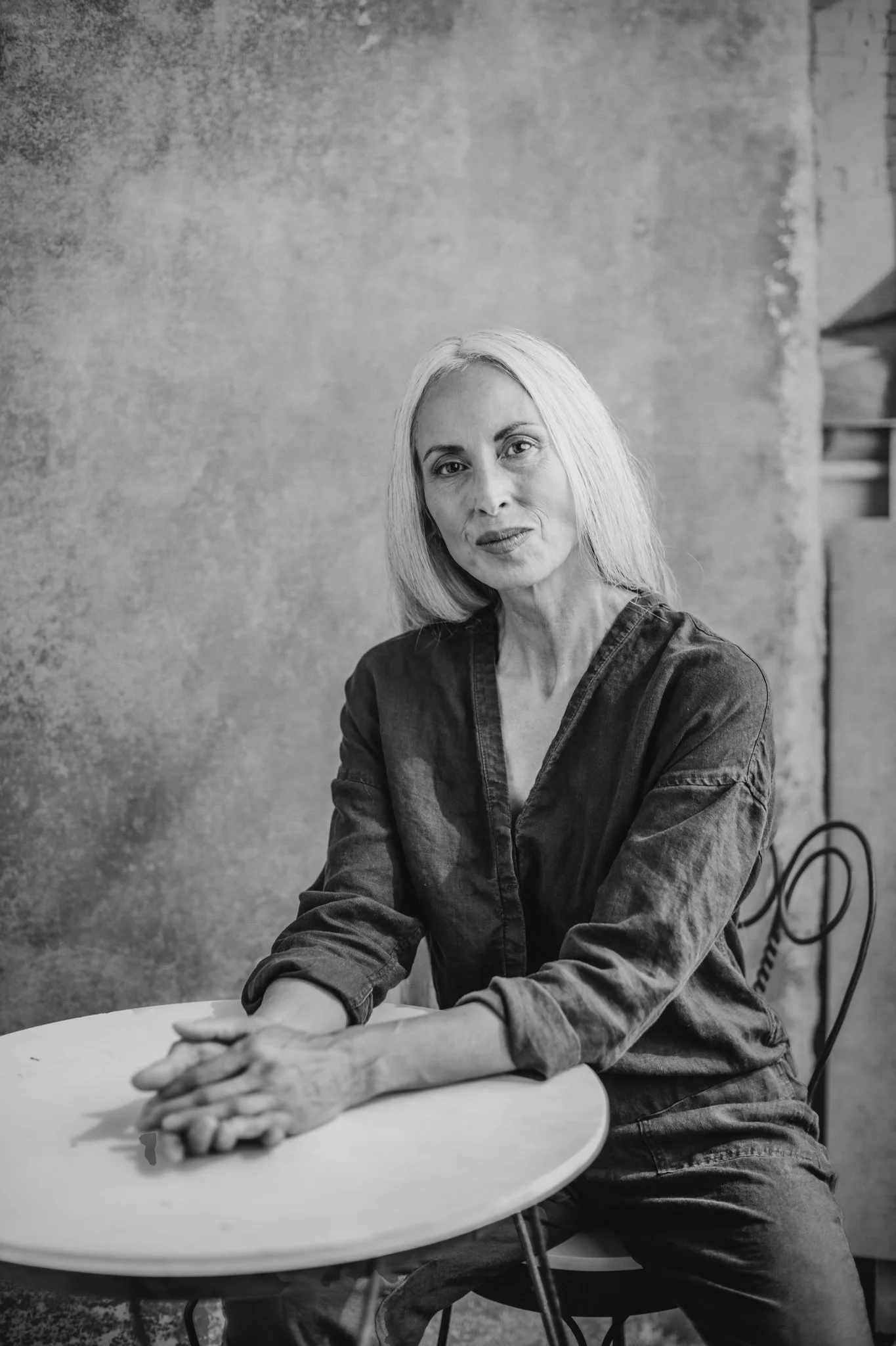 Black and white photo of a woman with light hair sitting at a table, looking at the camera with a slight smile, in an indoor setting with plain textured wall in the background.