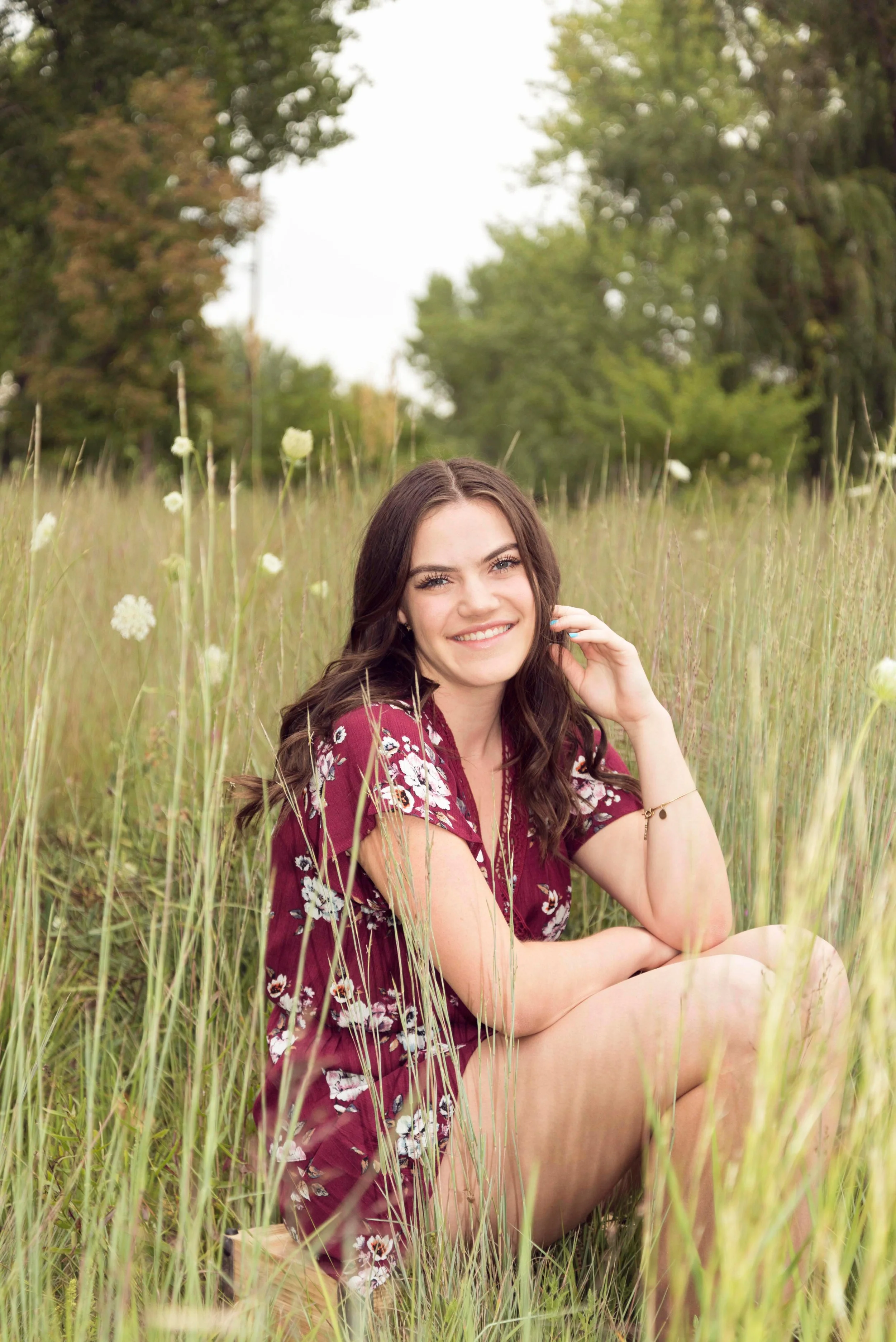 A young woman with long wavy hair, wearing a burgundy floral dress, sitting among tall grass and white flowers in a meadow with trees in the background, smiling at the camera.