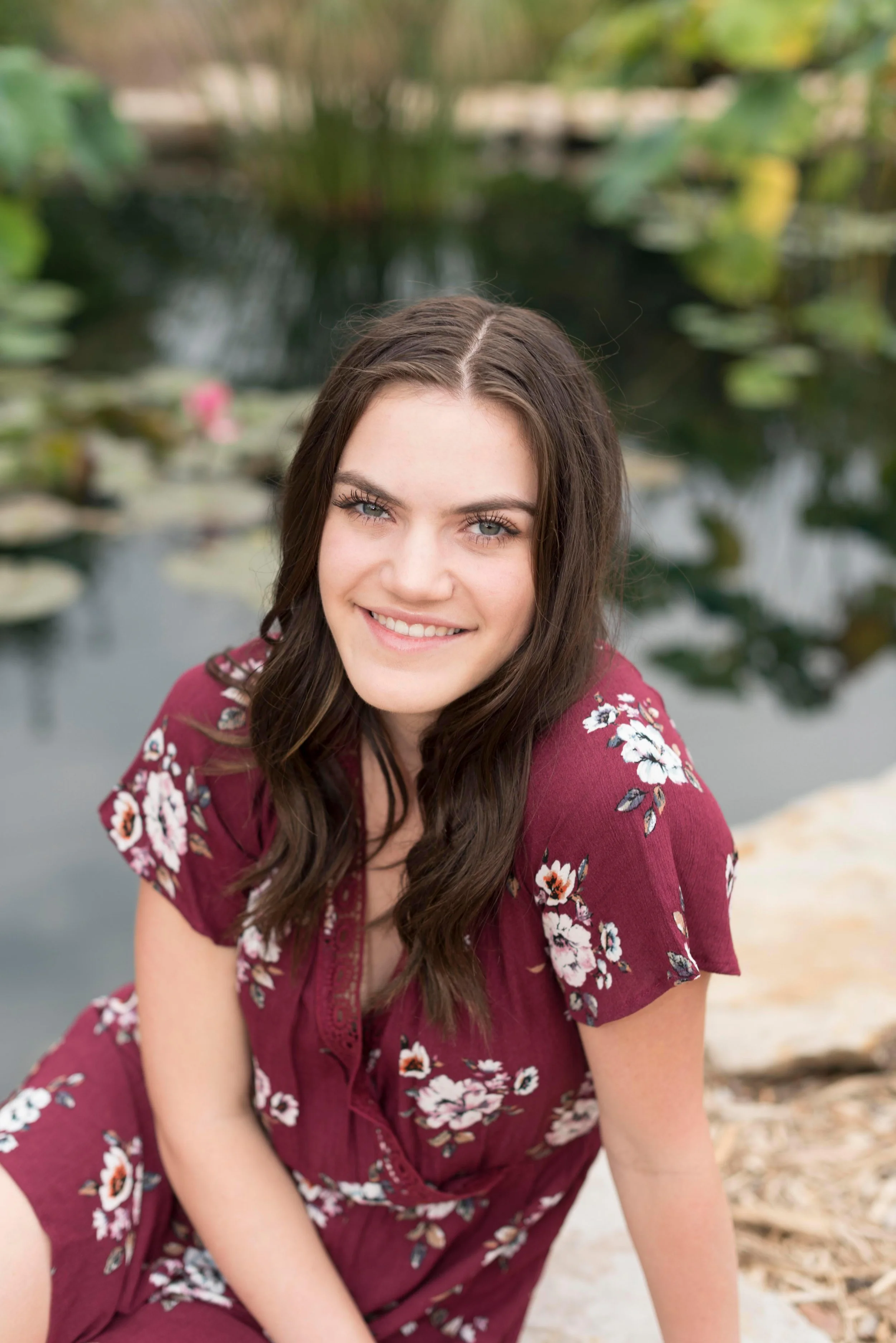 A young woman with long brown hair and blue eyes is smiling and sitting outdoors near a pond with lily pads and green plants.