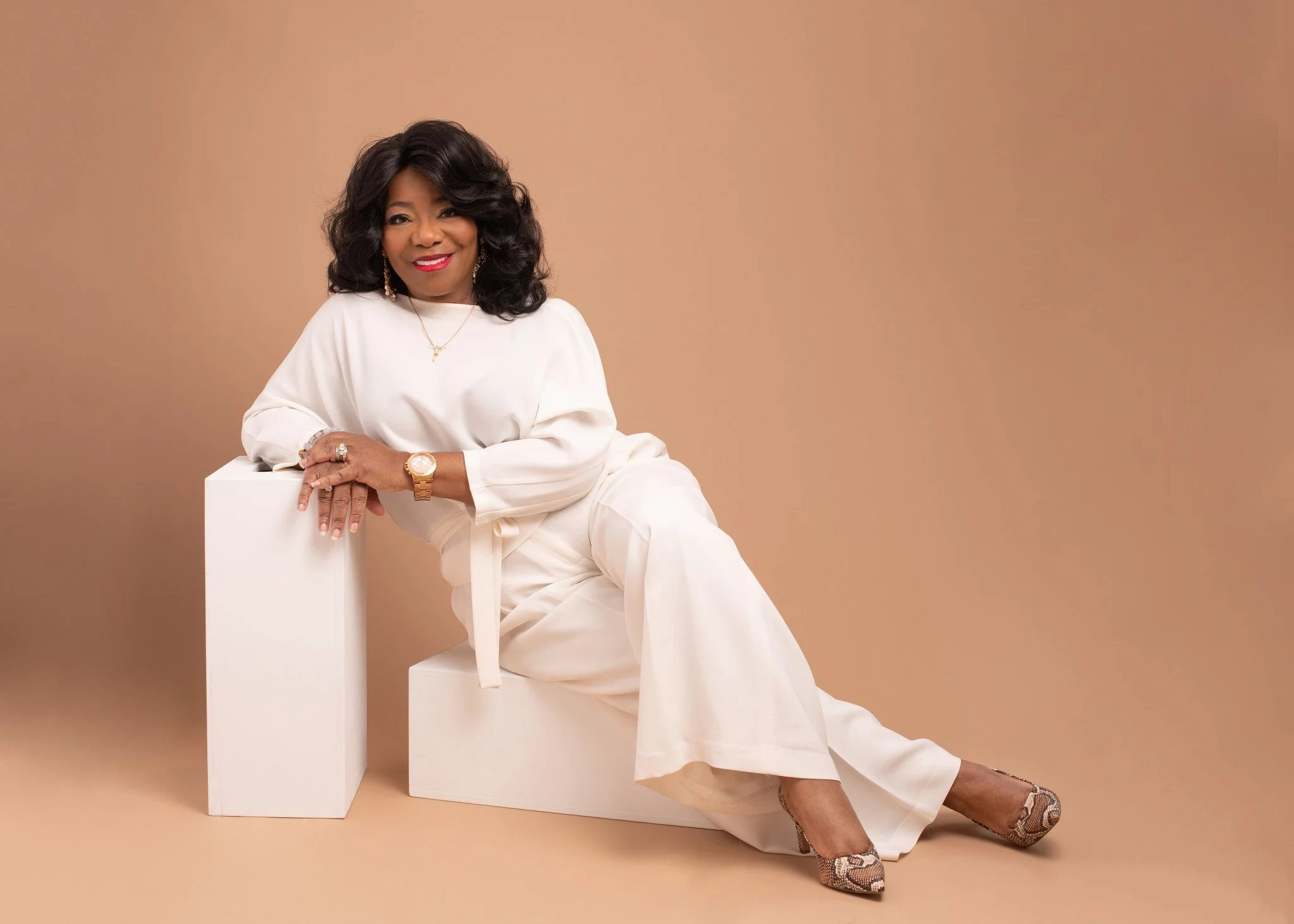 A woman with dark curly hair, smiling, dressed in white, sitting on a beige background. She is wearing gold jewelry, including a watch, necklace, earrings, and rings, and snakeskin patterned high heels.
