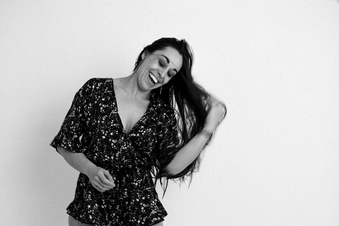 A woman with long hair in a floral dress laughing and brushing her hair back.