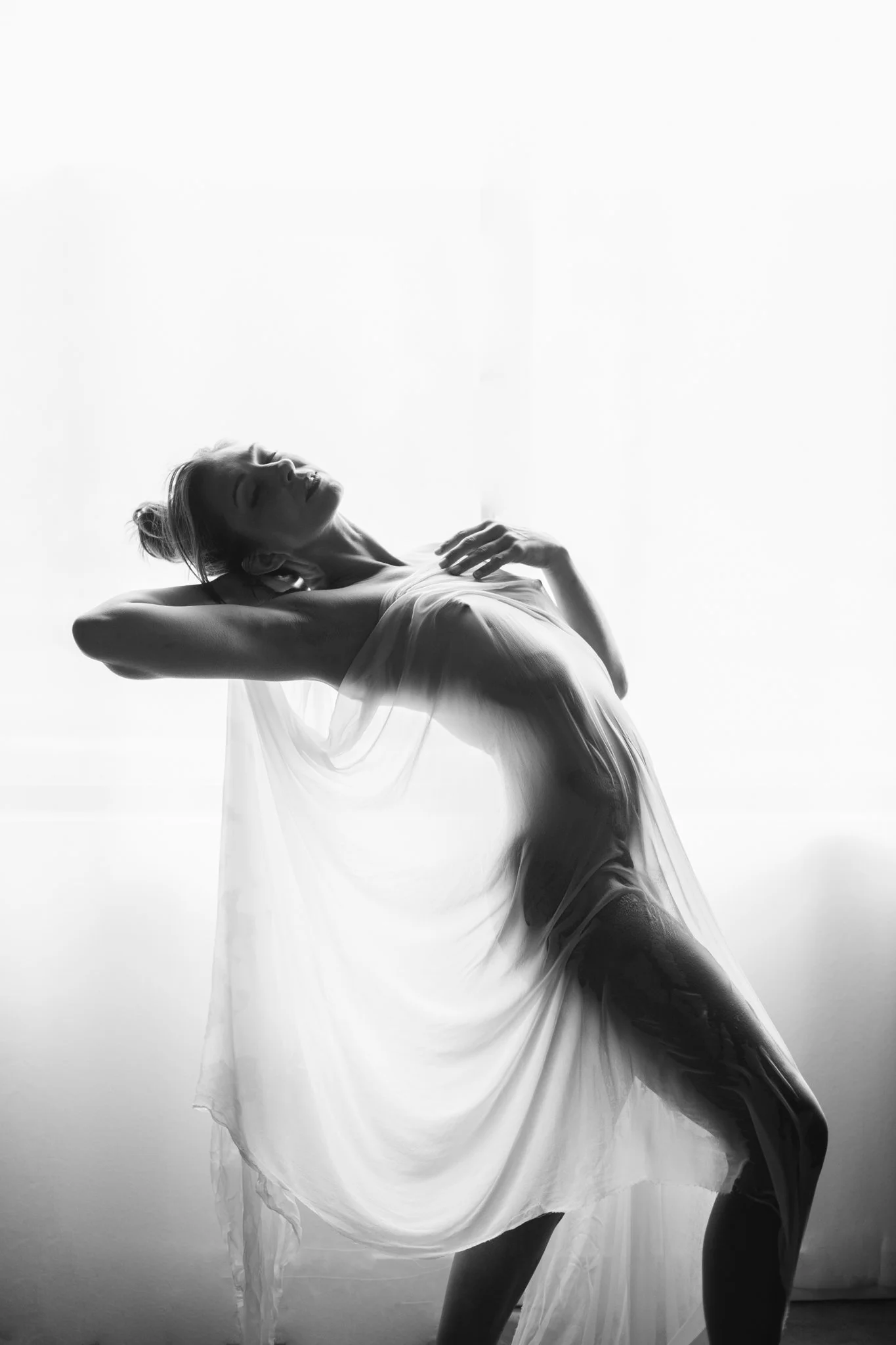 Black and white photograph of a woman with her eyes closed, wearing a flowing dress, with one arm behind her head and the other hand on her chest, standing against a bright background.