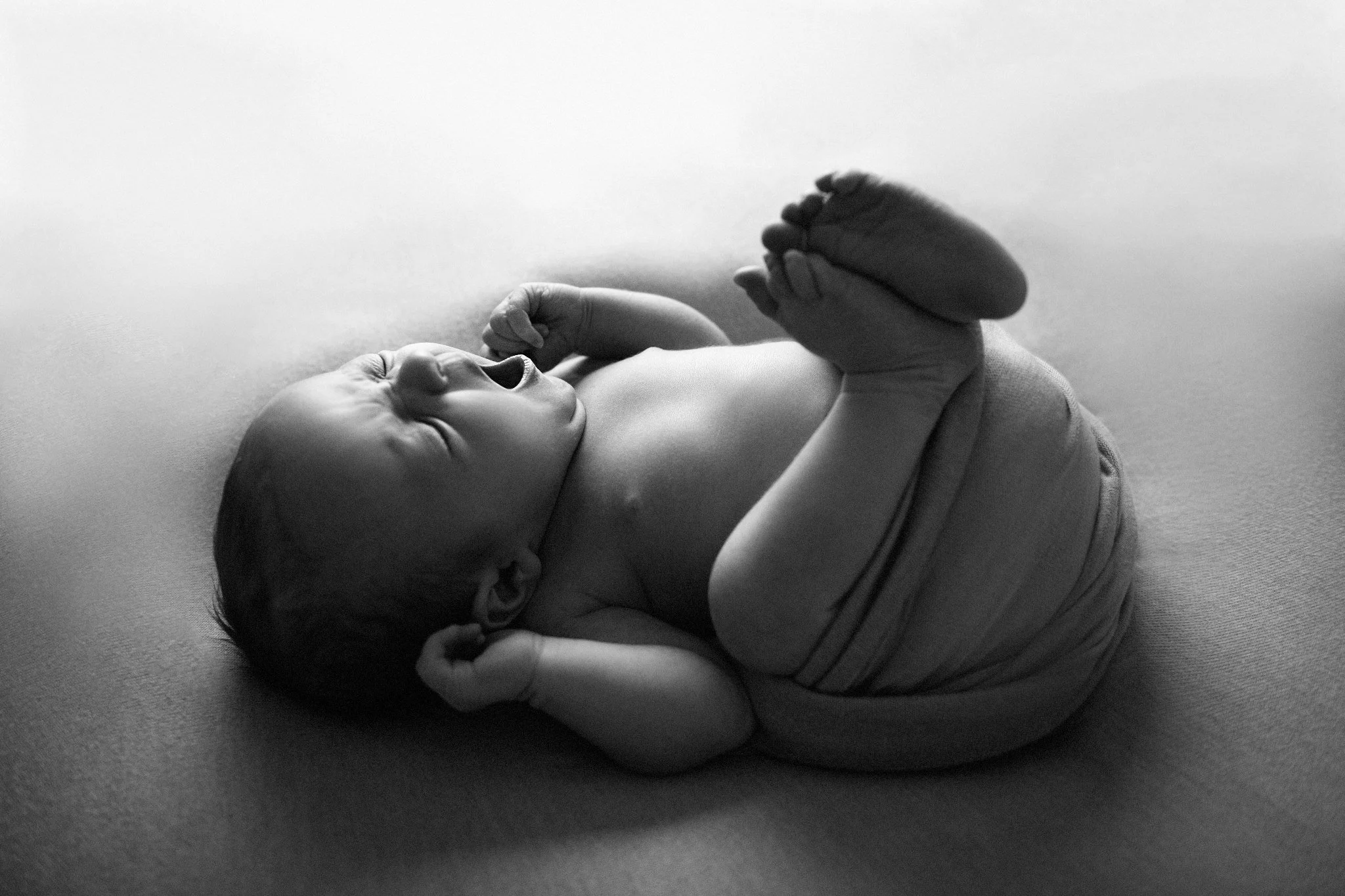 Black and white photo of a newborn baby lying on a blanket. The baby is smiling, with eyes closed, and holding one hand near its face.