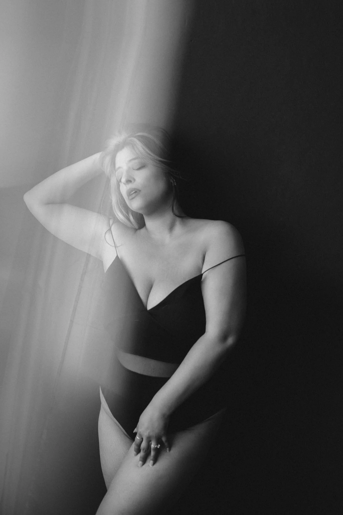 Black and white photograph of a woman posing with her eyes closed and head tilted back, wearing a strapless black top, with her right hand placed on her thigh and her left hand touching her hair.