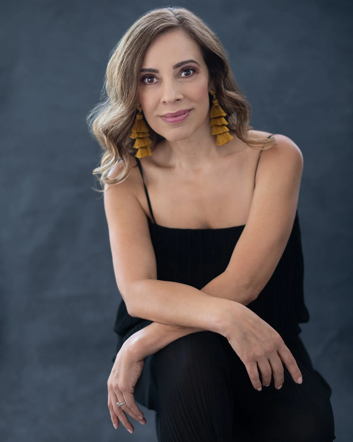 A woman with shoulder-length light brown hair wearing a black sleeveless dress and large yellow tassel earrings, sitting against a dark gray background.