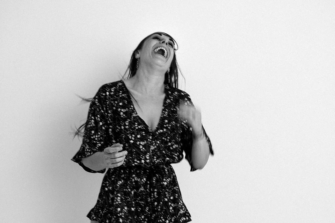 A woman with long hair wearing glasses and a patterned dress, laughing with her head tilted back.