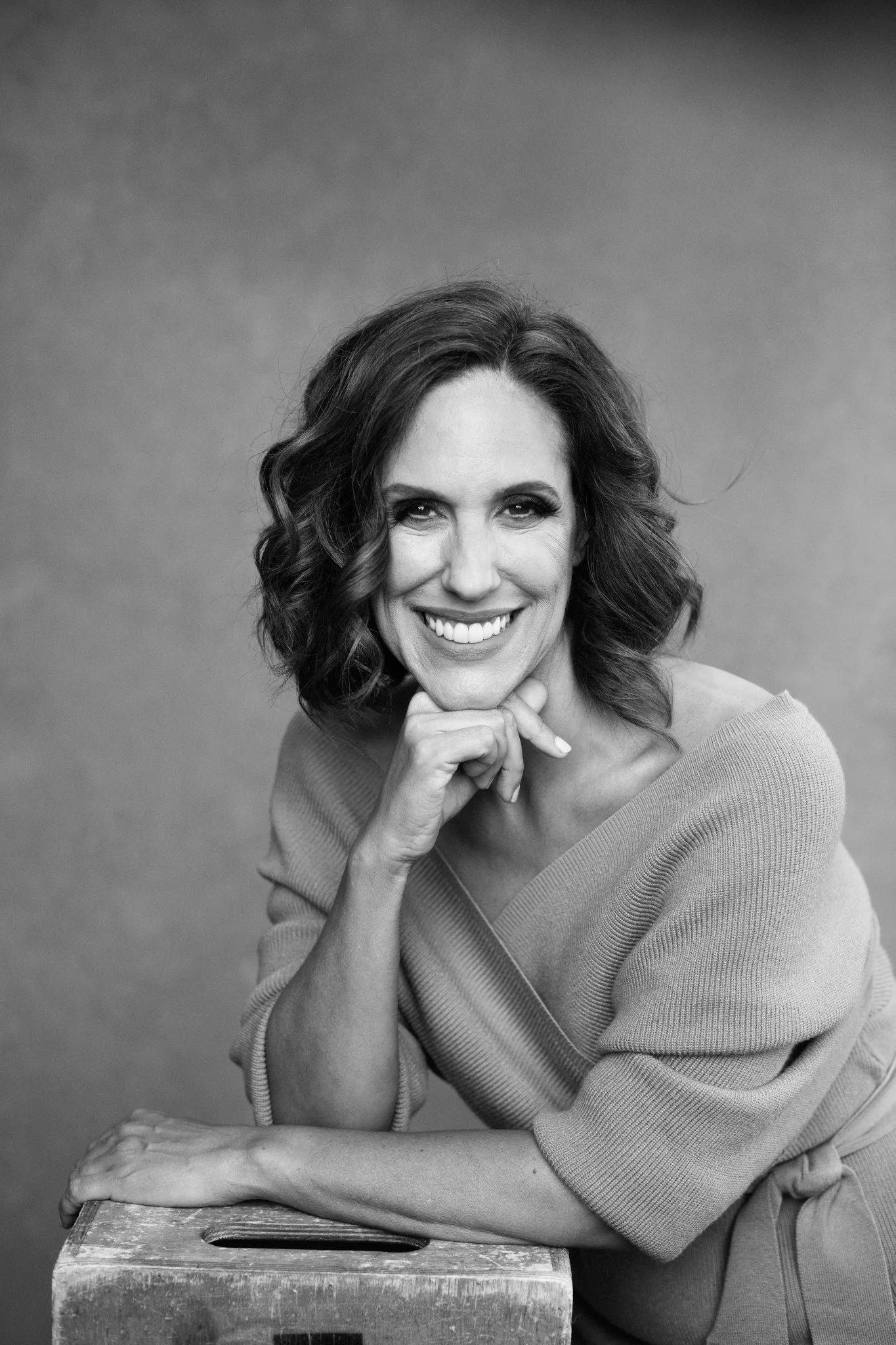 Black and white portrait of a woman smiling, with her chin resting on her hand, sitting next to a wooden box, against a plain background.