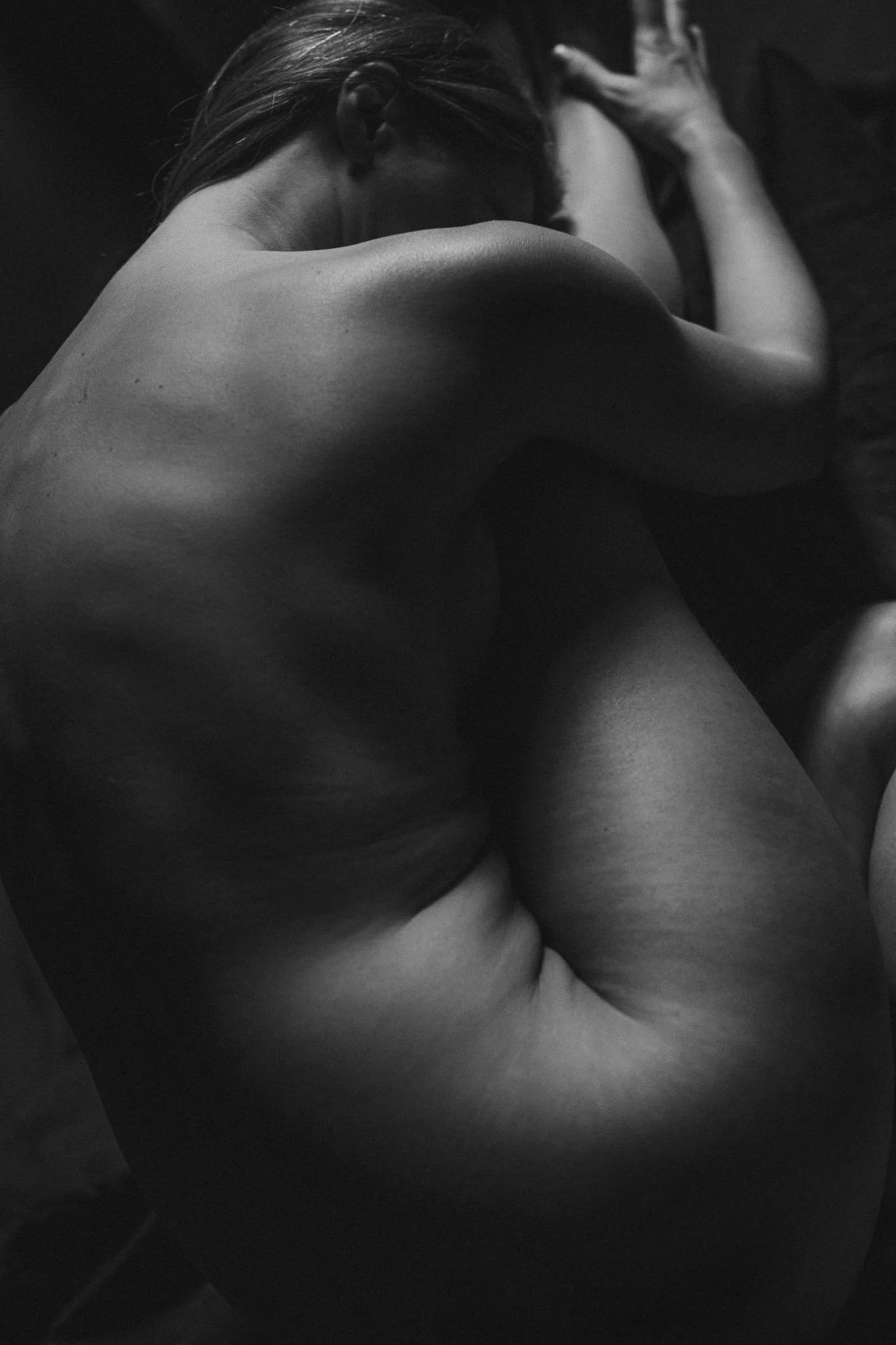 Black and white photograph of a nude person curled up on a dark surface, with their head resting on their arms.