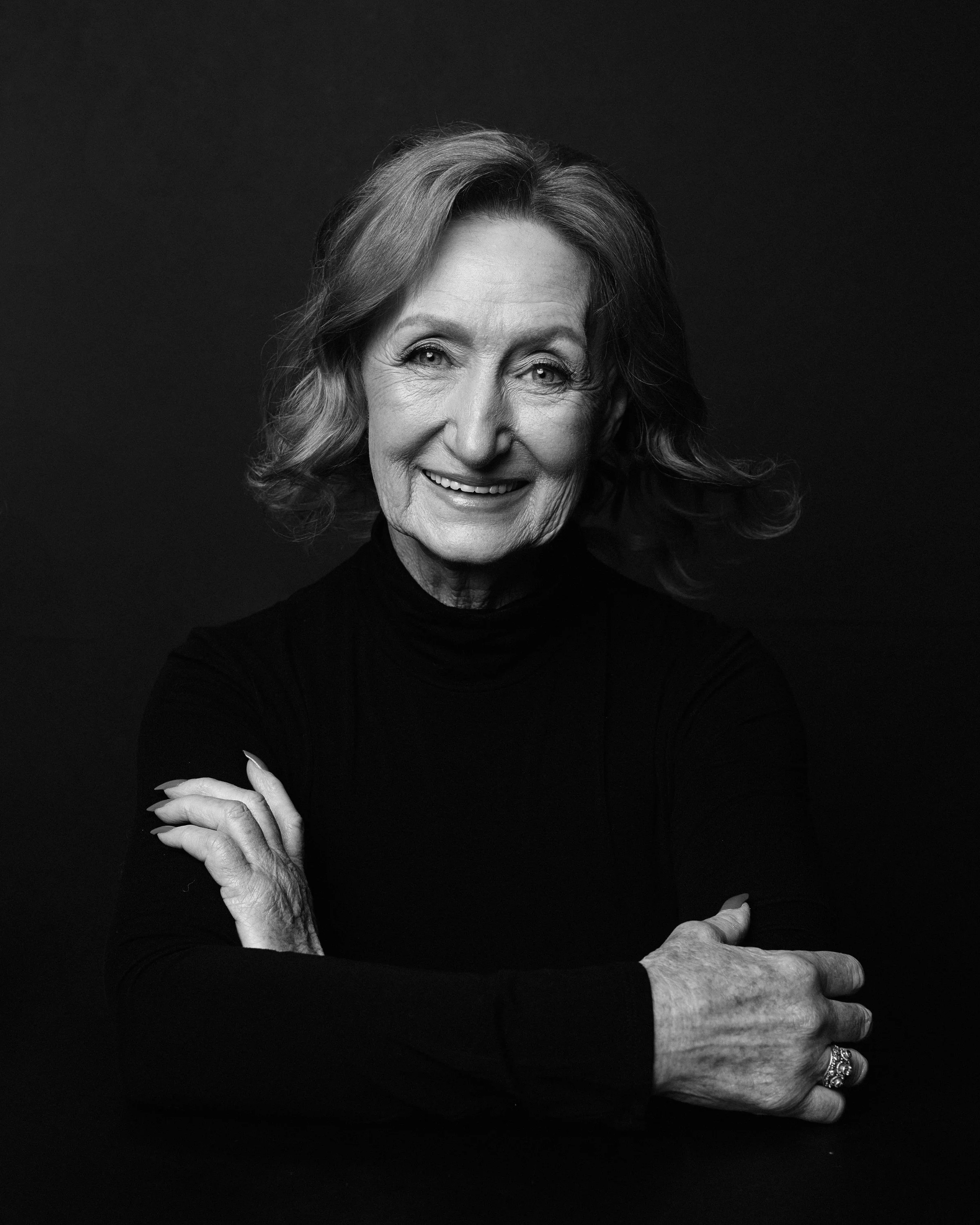 Black and white photograph of an older woman with short, wavy hair smiling, wearing a dark top, with her arms crossed. An elderly person's hand rests on her arm, and another person's hand is in view at the bottom right corner.