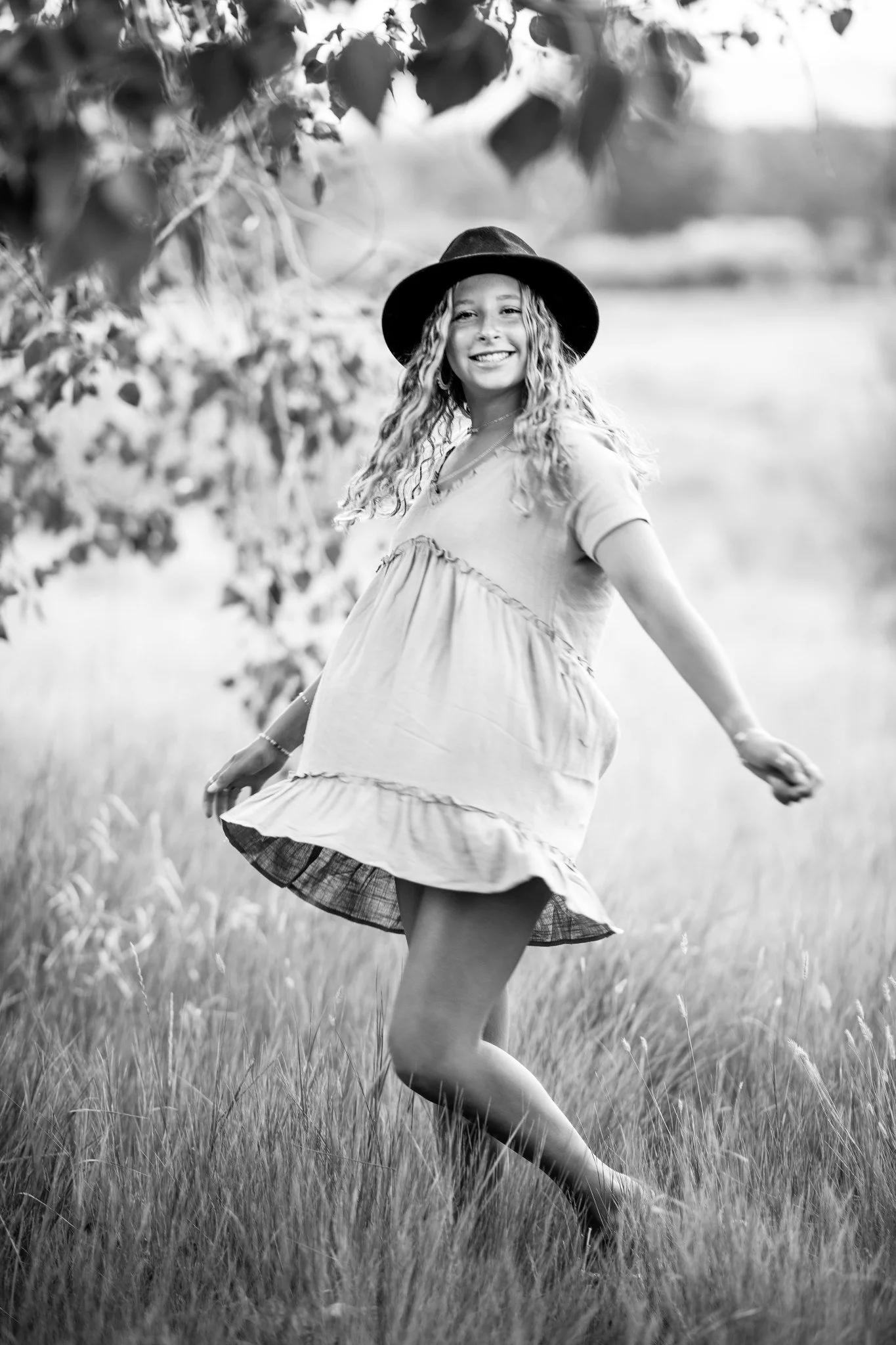A smiling young woman with curly hair wearing a wide-brimmed hat and a loose dress, dancing in a grassy field with trees and a body of water in the background.