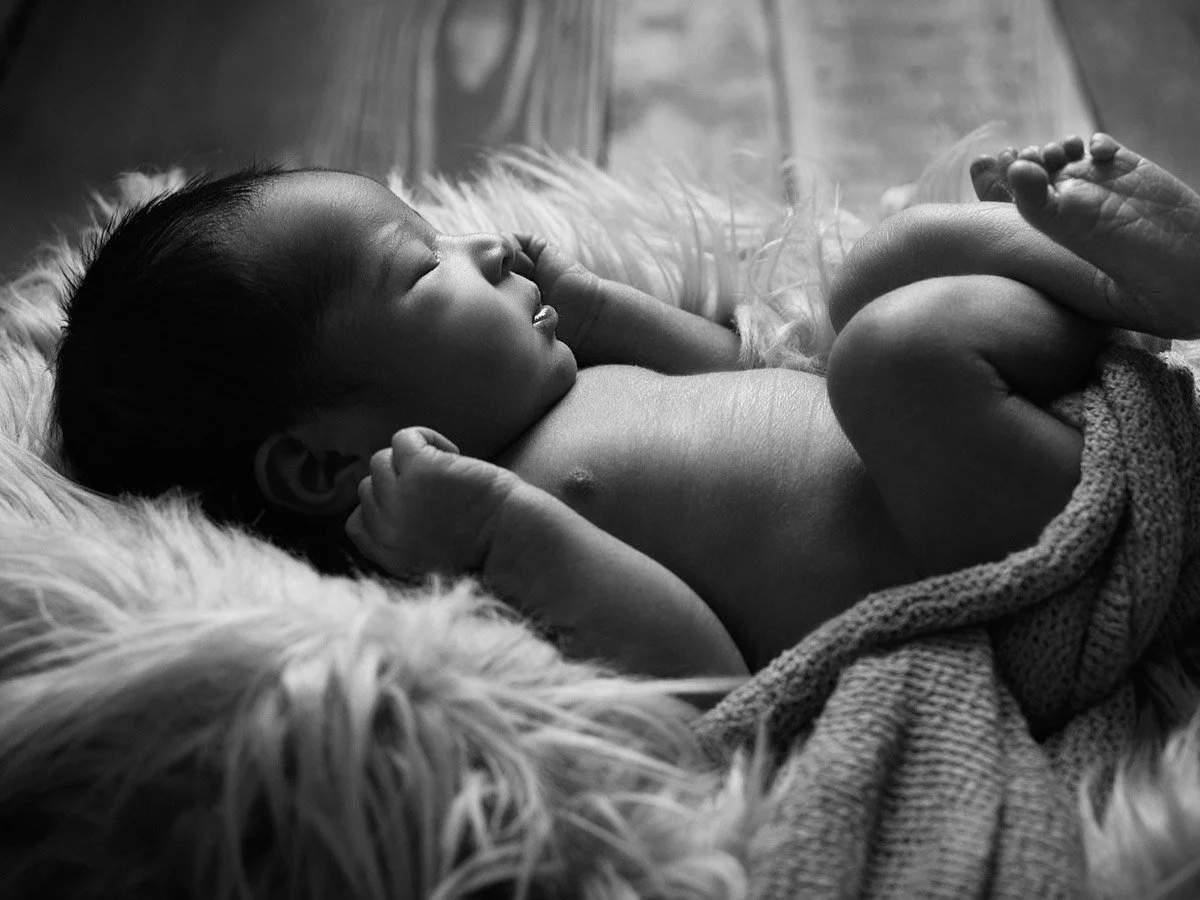 A sleeping baby lying on a soft, furry surface with a blanket draped over their lower body.