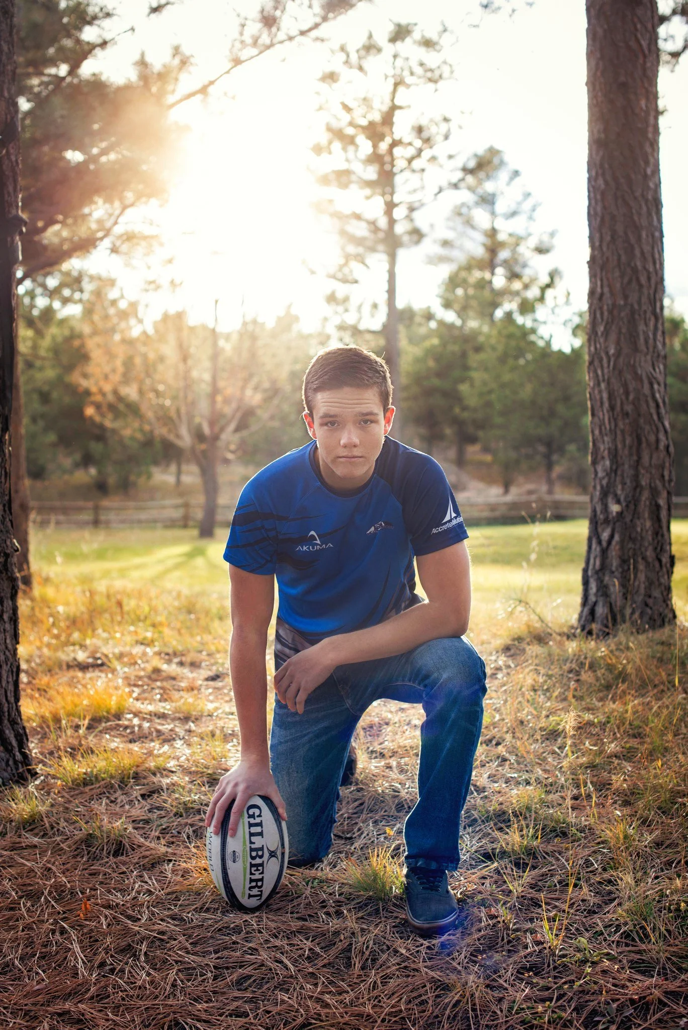 A young man in a blue sports shirt kneels on one knee on a forest floor, holding a rugby ball, with trees and sunlight in the background.