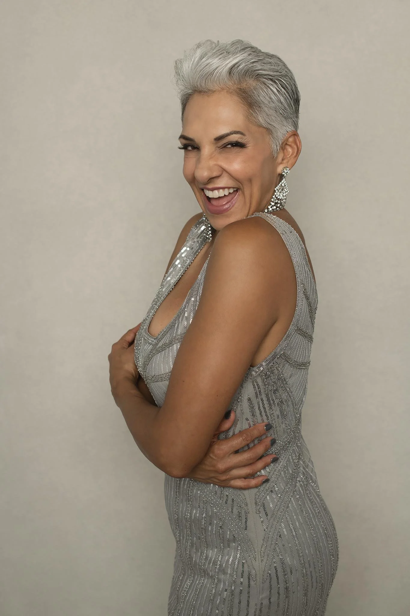 A woman with short gray hair wearing a silver, sequined gown and earrings, smiling and posing with her arms crossed against a plain background.