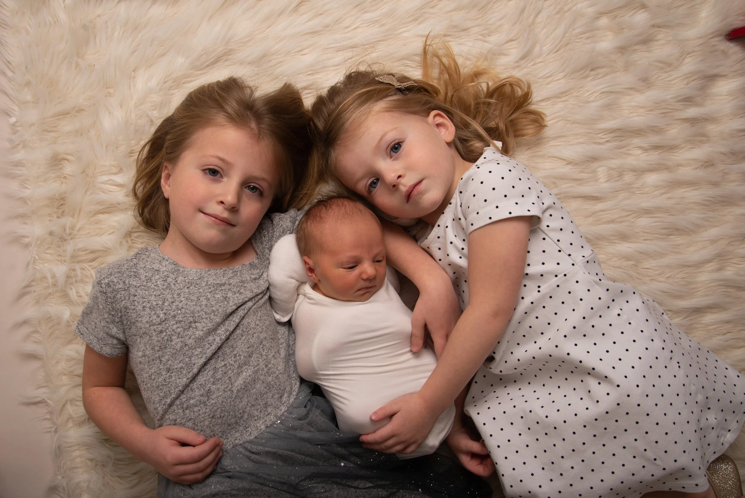 Four children lying down on a fluffy white rug, with their heads close together. Two older girls and a baby, all looking at the camera.