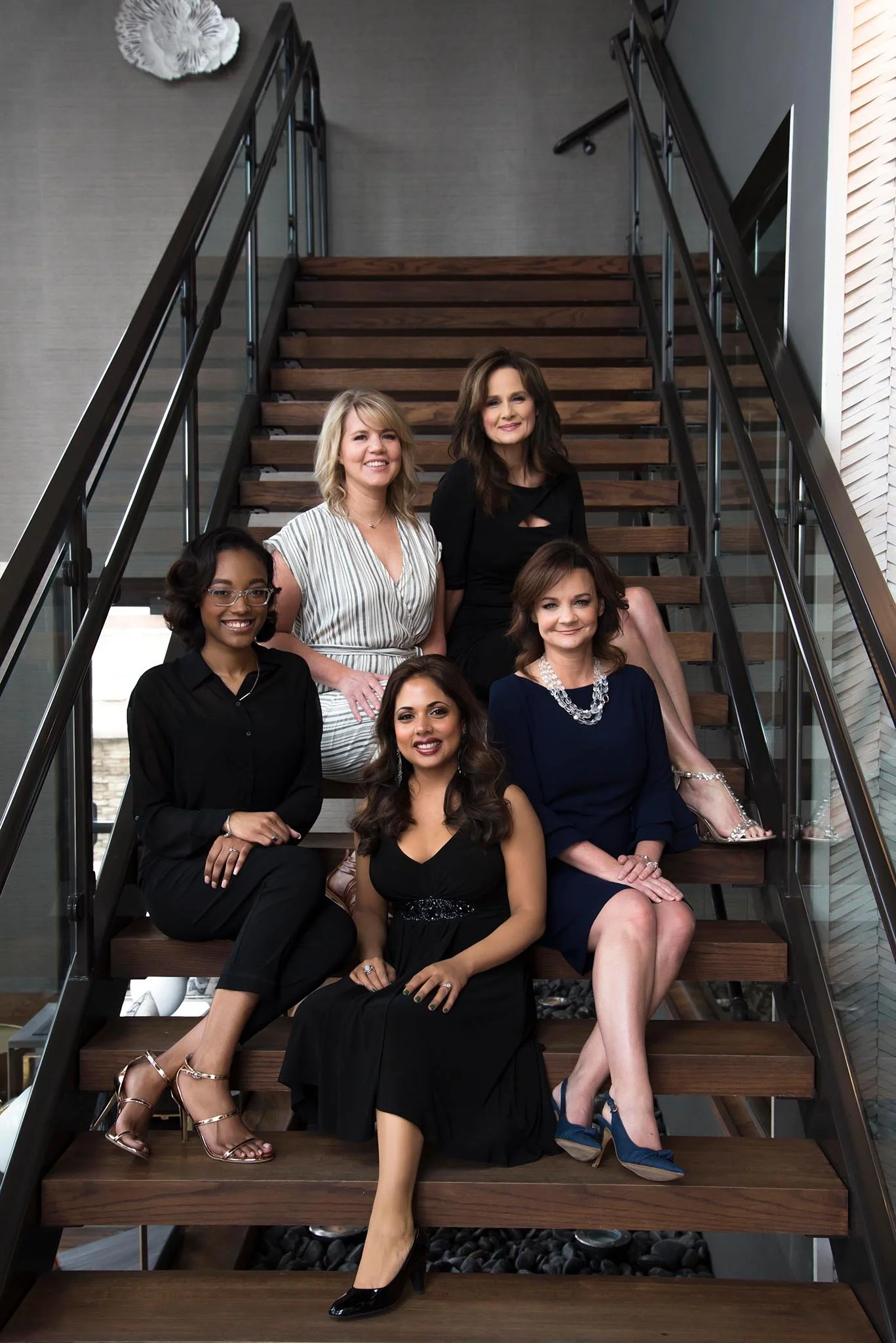 Group of six women sitting on a modern staircase, dressed in formal attire, smiling for the camera.