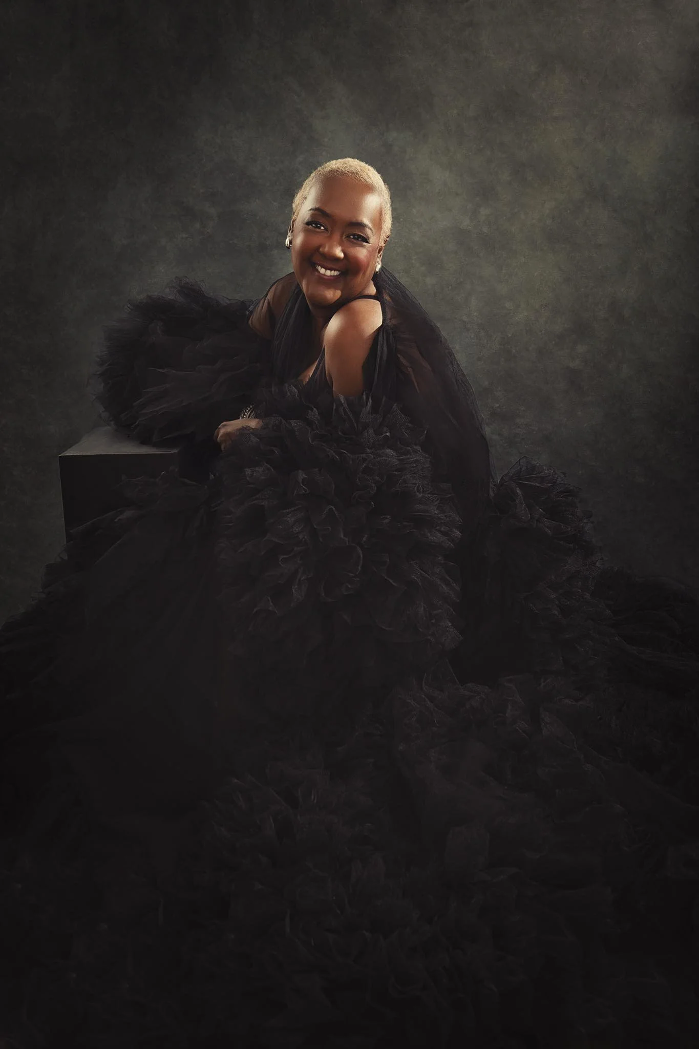 A woman in a black dress with ruffled fabric, smiling and leaning on a black platform against a dark textured background.