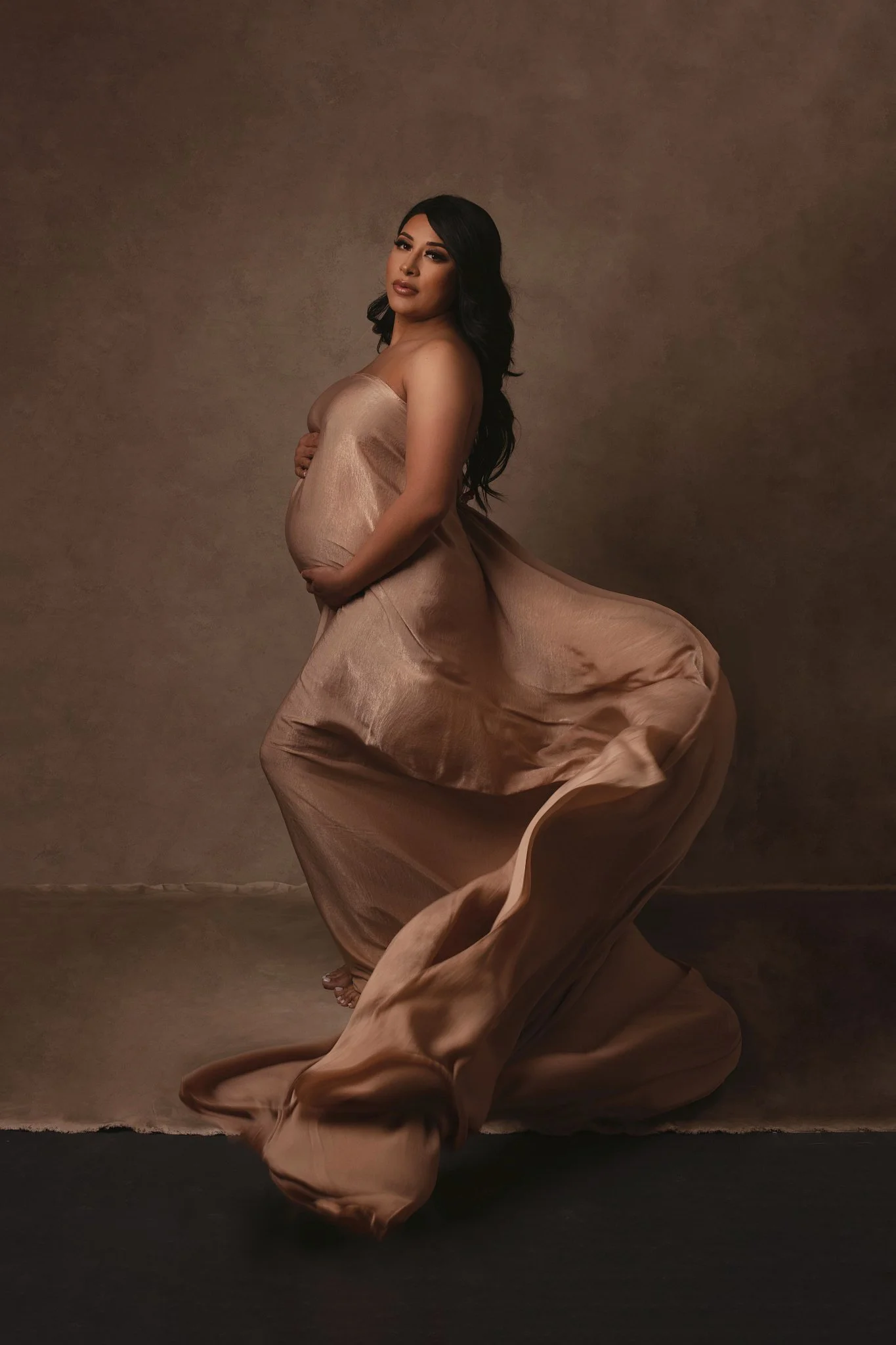 A pregnant woman with long dark hair, wearing a flowing satin dress, standing against a plain brown background.