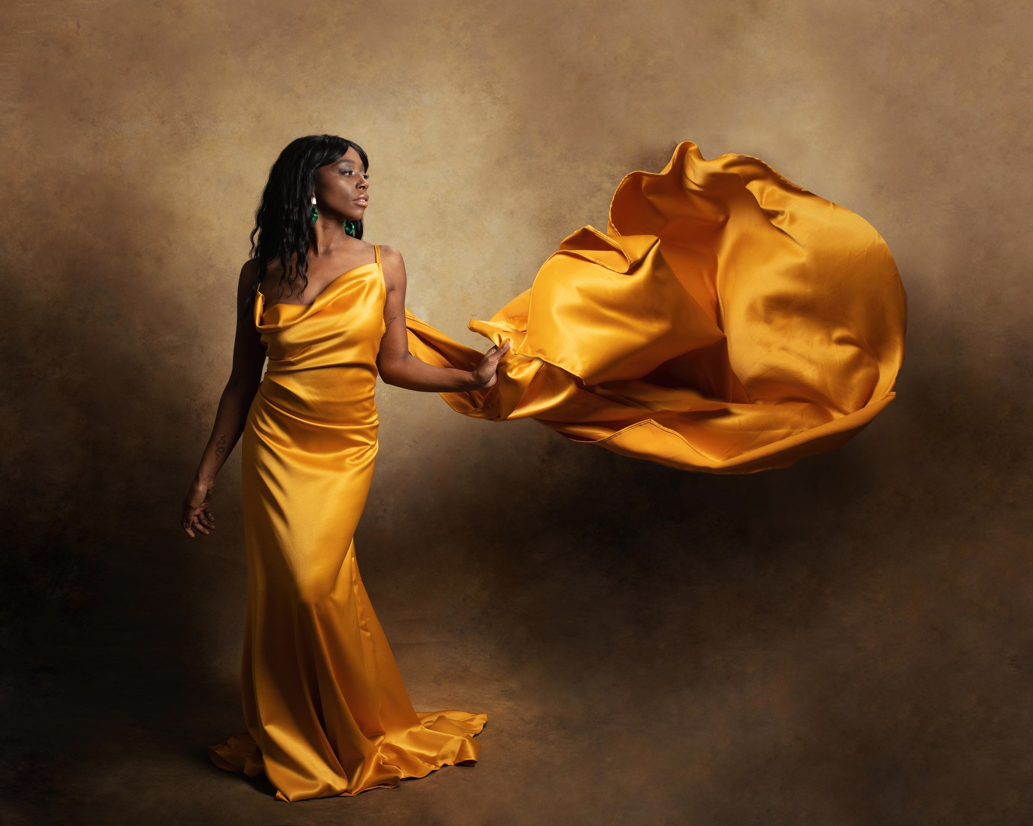A woman in a yellow satin gown holding the fabric of her dress, the background is a textured gradient in brown tones.