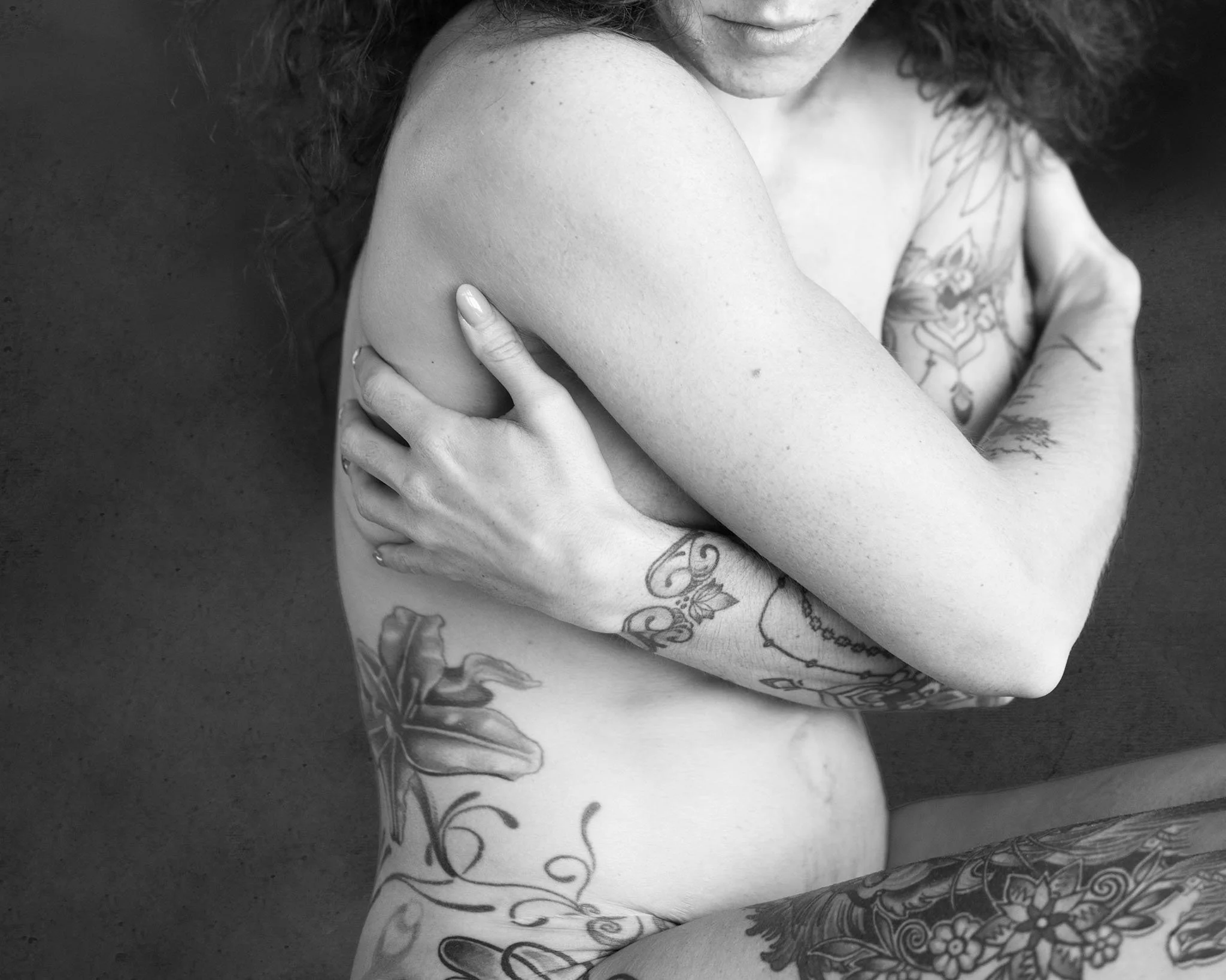 A black and white photo of a woman with tattoos, partially sitting and hugging herself, with her arms crossed over her chest, showing her tattoos on her arms, torso, and legs.