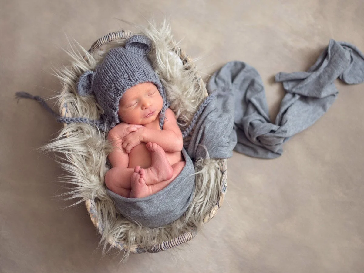 A sleeping newborn baby dressed in gray, wearing a knit hat with bear ears, and laying in a basket with furry lining and gray clothes draped around. The basket is on a beige surface.
