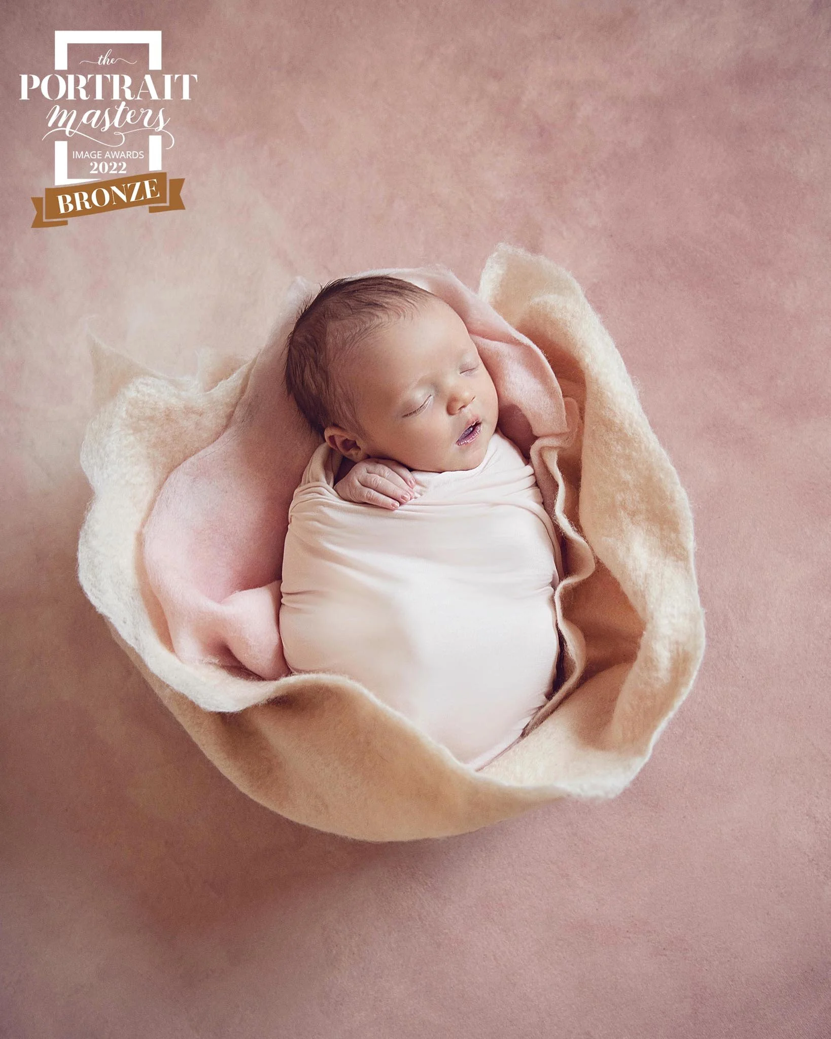 A sleeping baby wrapped in a light pink blanket, lying in a soft, beige, petal-shaped nest on a pink background.
