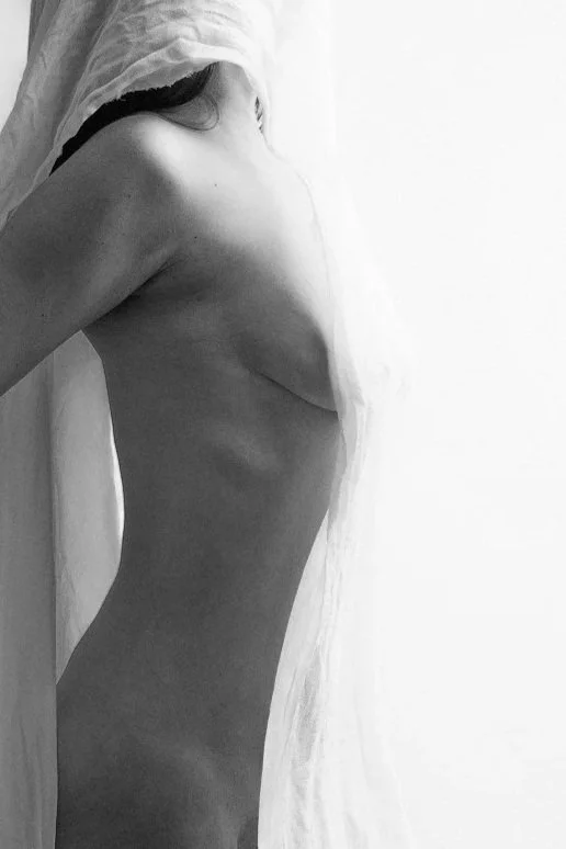 A black and white photo of a topless woman with a sheer cloth covering part of her head and face, standing next to a white curtain.