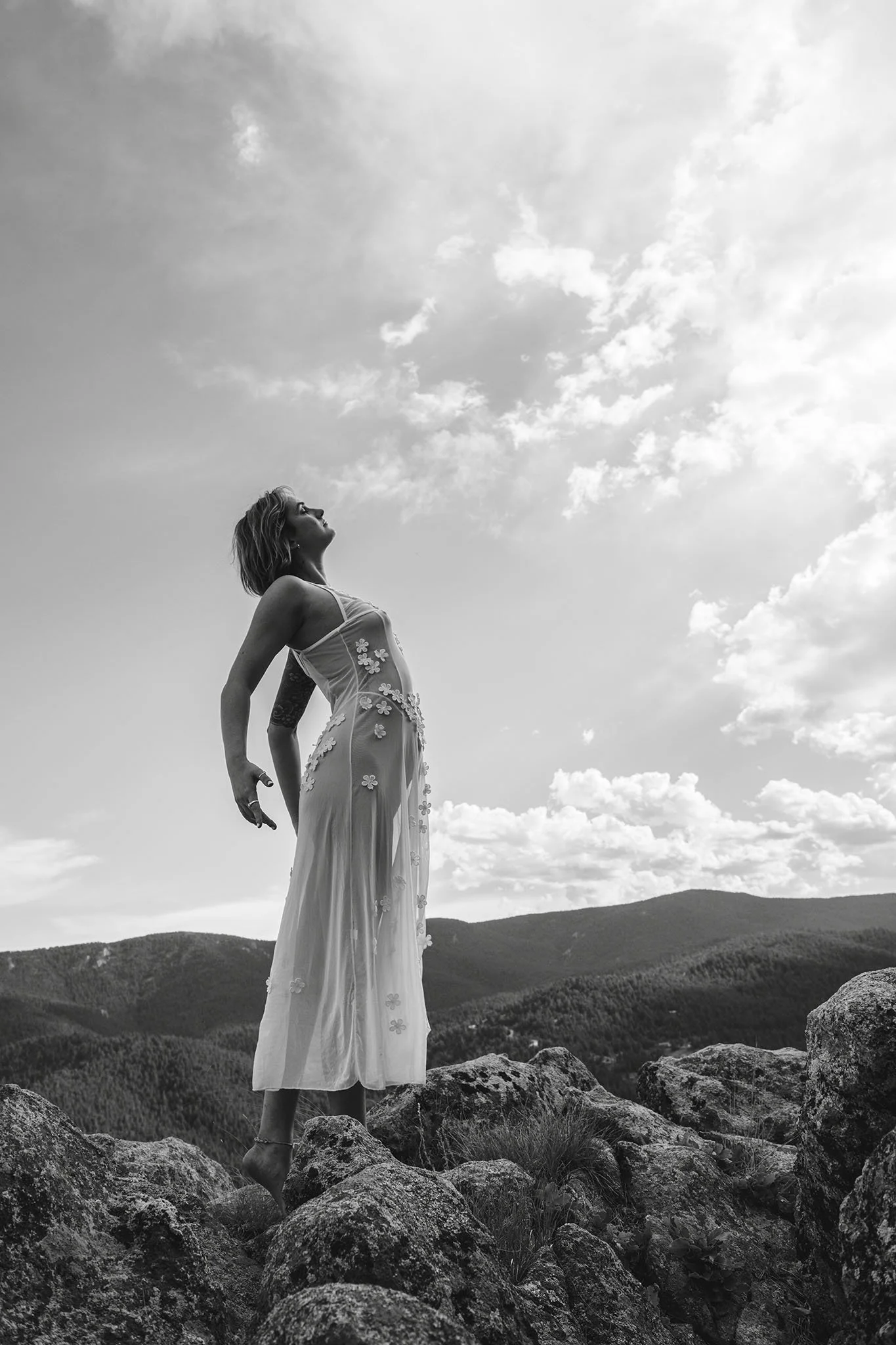 A woman standing on rocks in a scenic outdoor landscape, looking up at the sky with clouds and rolling hills in the background, wearing a flowy dress with floral details.