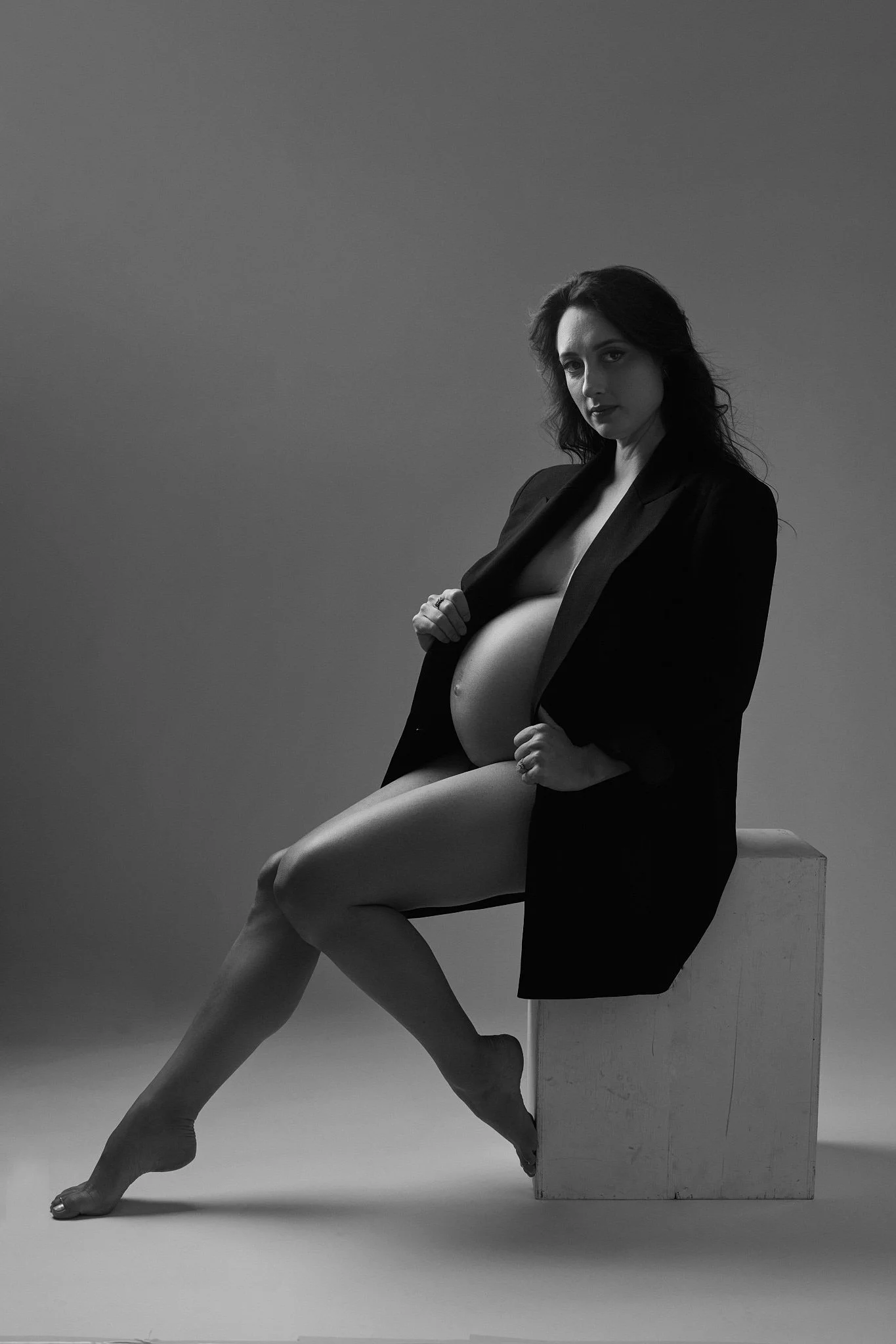 Black and white photo of a woman sitting on a chair with her legs crossed, wearing a blazer, revealing her thigh, with a plain gray background.