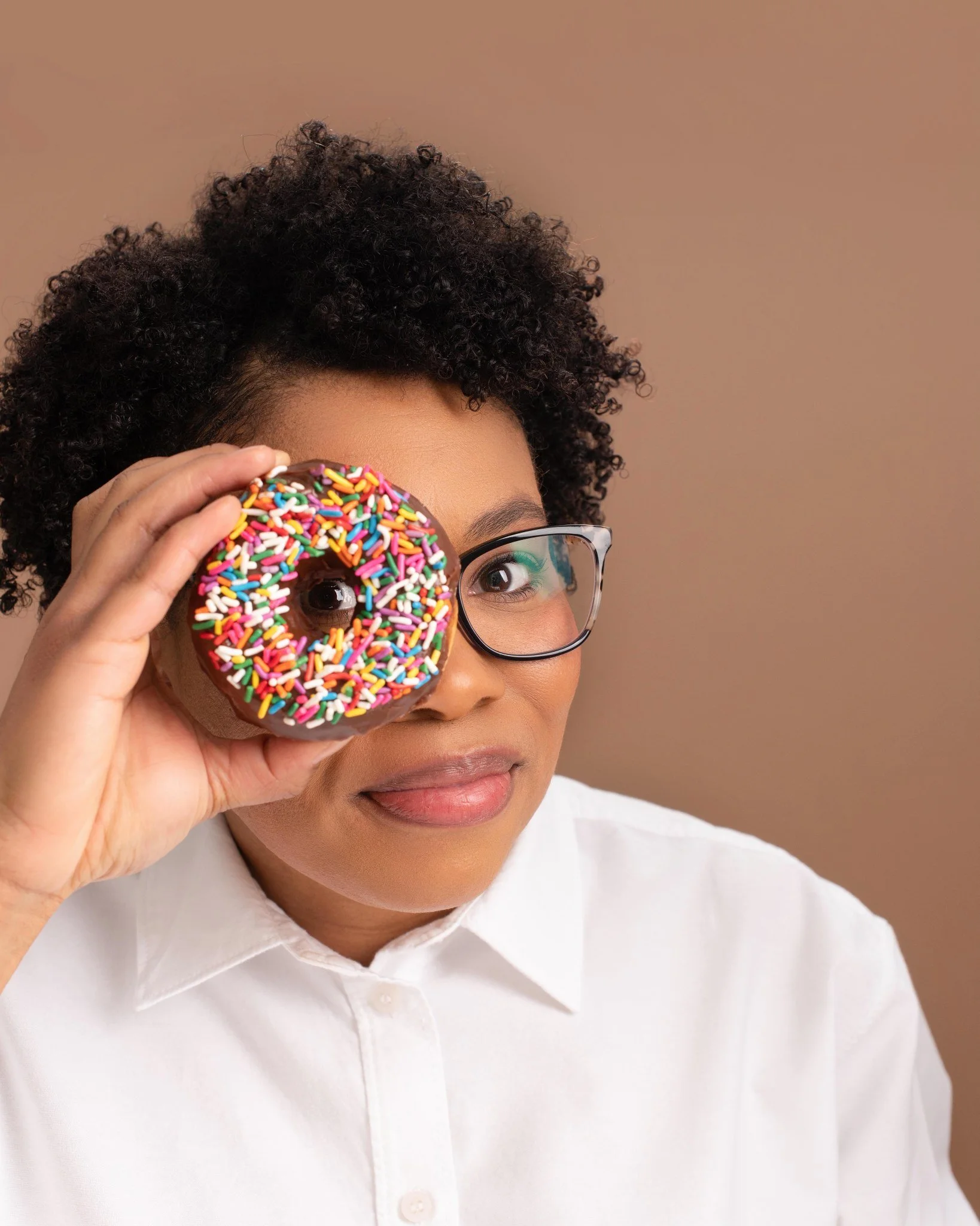 A woman with short curly hair and glasses holding a colorful sprinkled donut up to her eye.