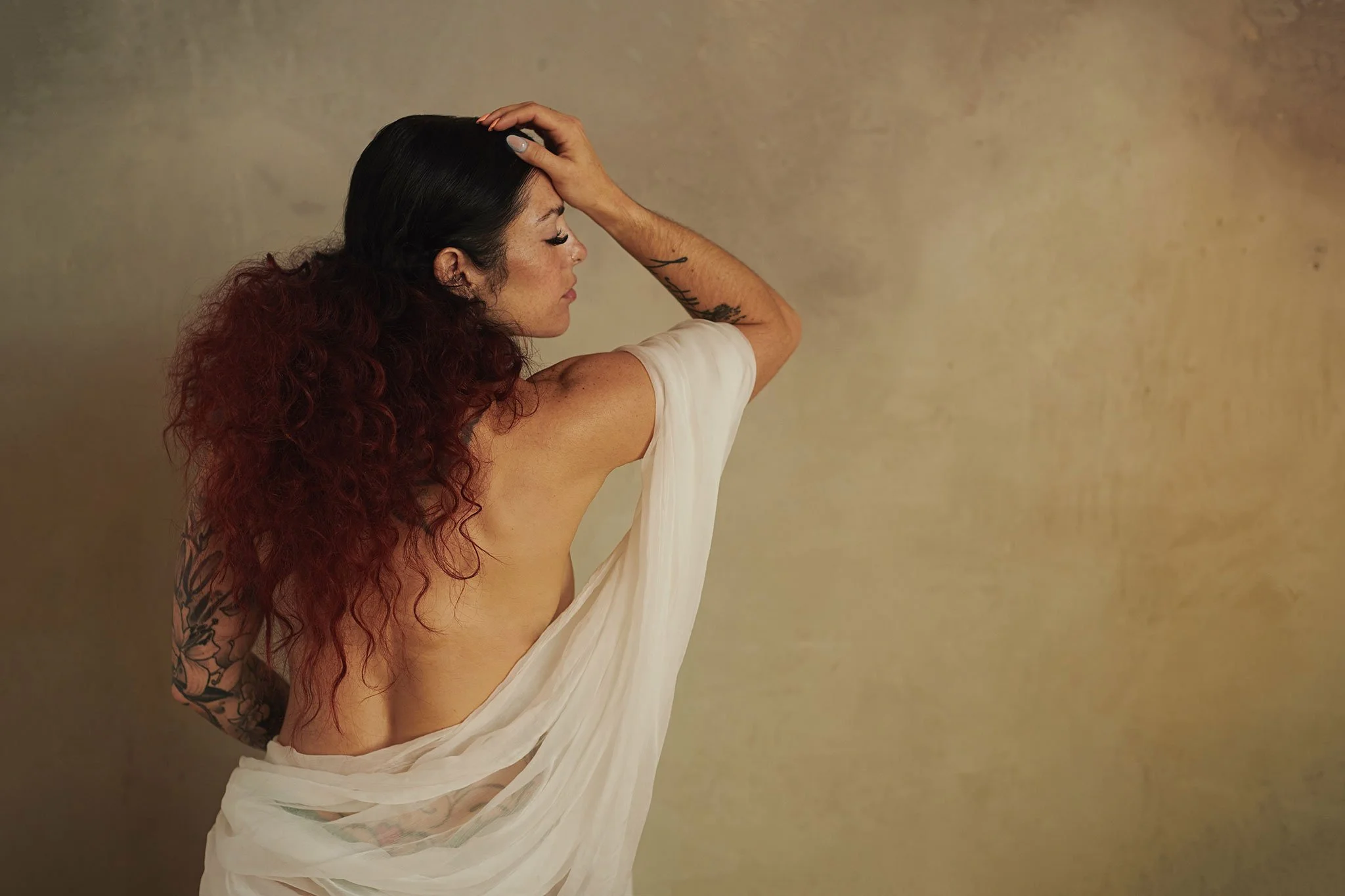 A woman with long, curly dark red hair and tattoos on her left arm poses with one hand on her head and eyes closed, wearing a white fabric draped over her shoulder against a plain beige background.