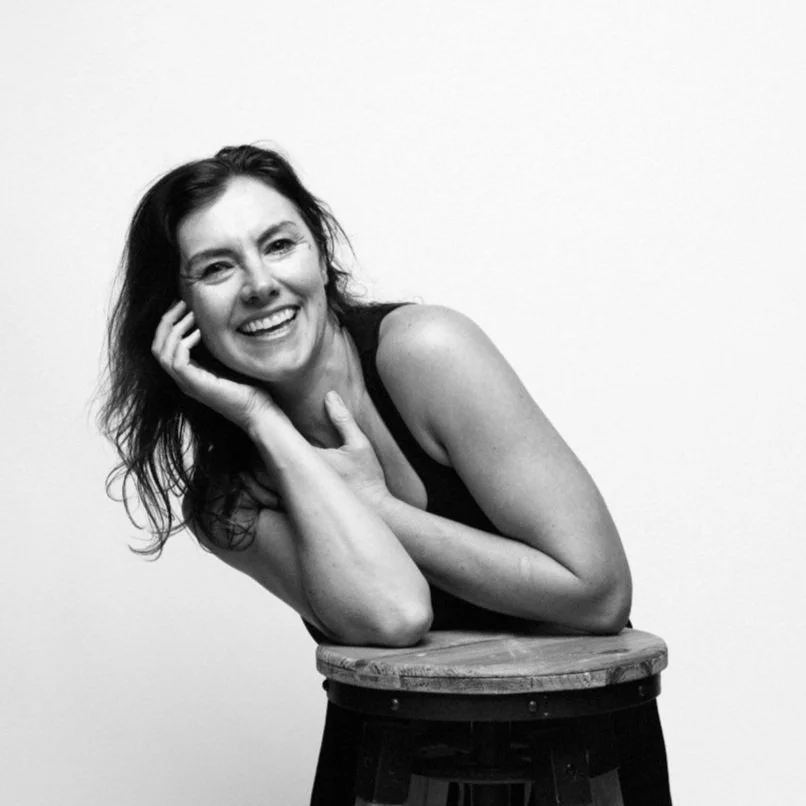 Black and white photo of a woman with wavy hair, smiling, resting her head on her hand, leaning on a wooden stool.