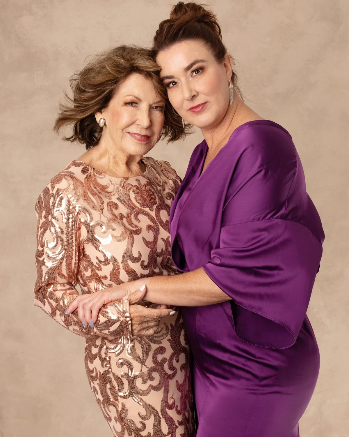 Two women, one older and one younger, dressed elegantly, are posing together, holding hands and looking at the camera against a plain background.