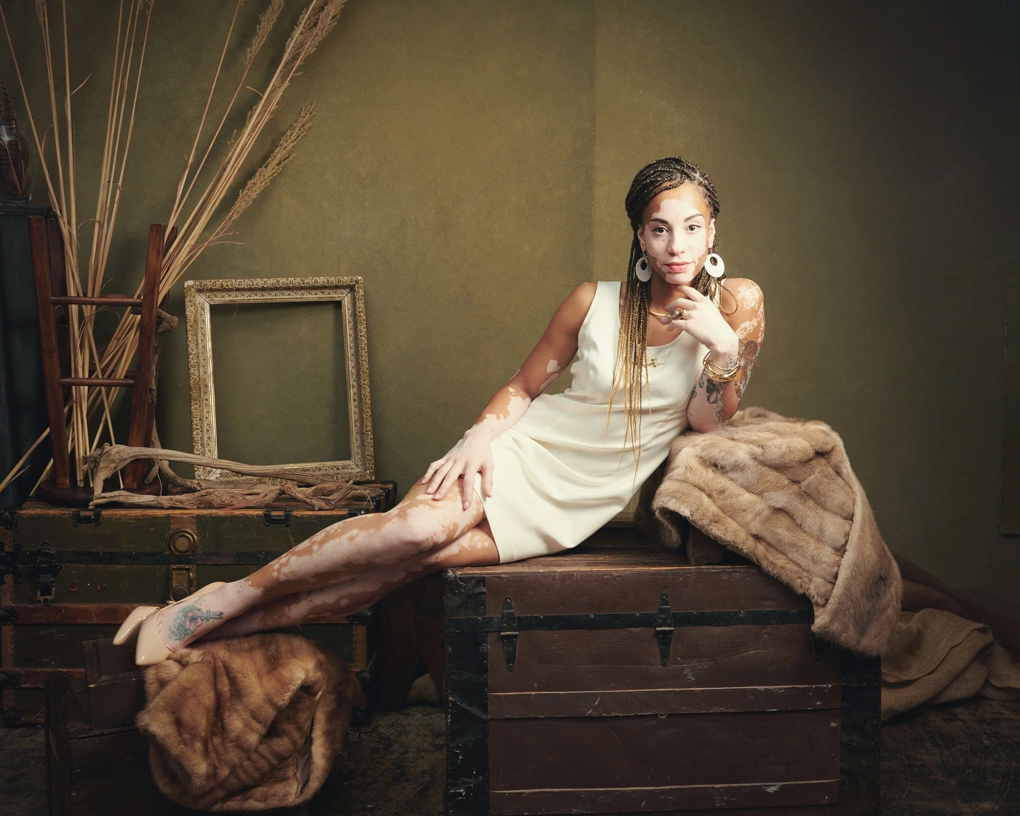 A woman lying on a vintage wooden chest with a fur coat, in a room decorated with dried plants, empty picture frames, and a green wall.