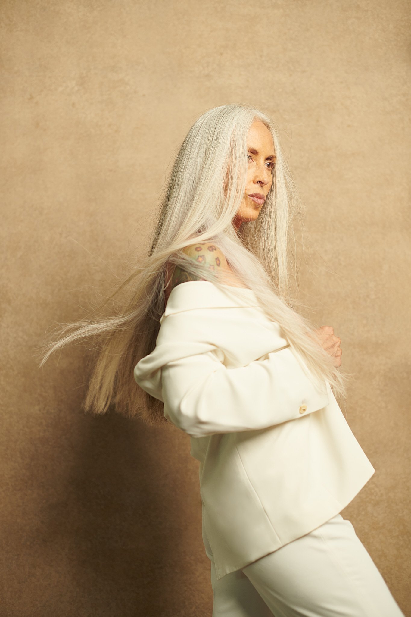 A woman with long, straight, platinum blonde hair and tattoos on her shoulder, wearing a white off-shoulder jacket and matching white pants, standing against a beige background.
