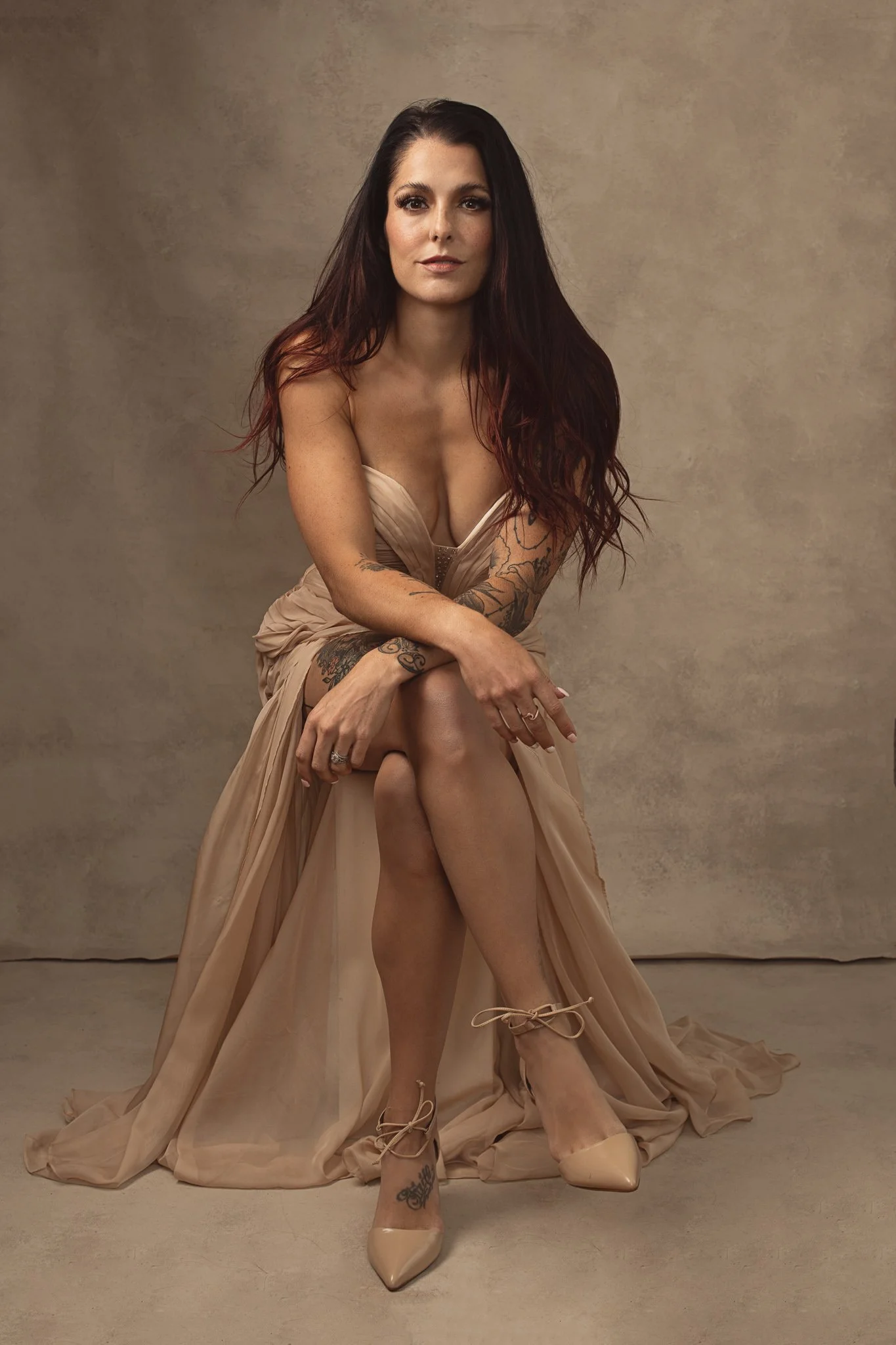A woman with long dark hair, wearing a beige dress, sitting with one leg crossed over the other, in front of a plain beige background.