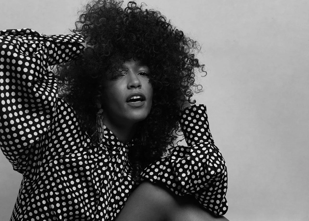 Black and white photo of a woman with curly hair wearing a polka dot blouse, sitting against a plain background.