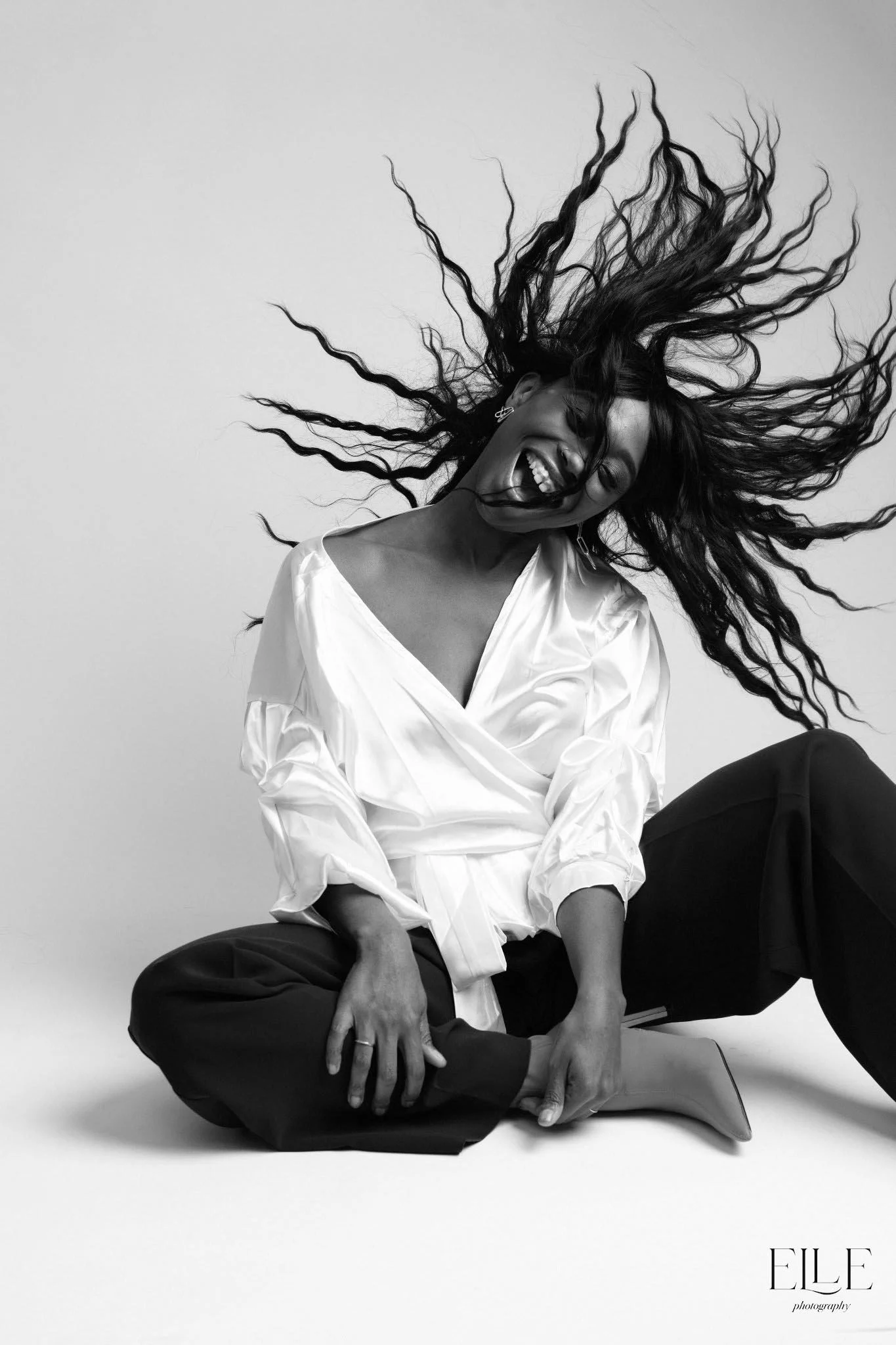 A woman with long, wavy hair spinning and smiling, sitting on the floor with her legs crossed, wearing a white wrap blouse and dark pants, against a plain background.