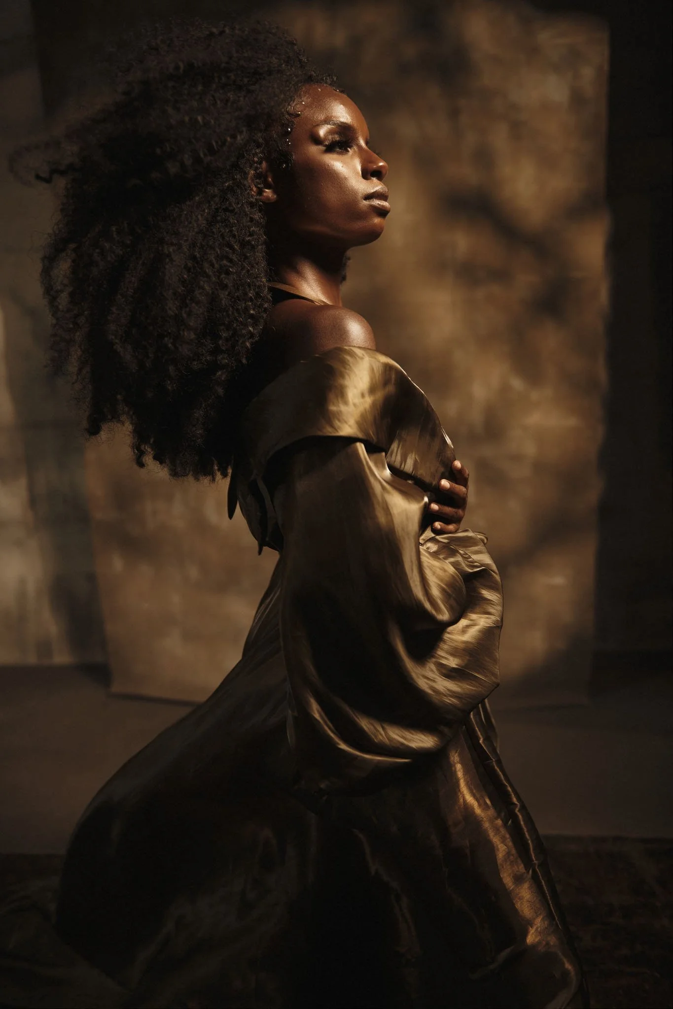 A woman with dark skin and curly hair, wearing an oversized brown satin coat, kneeling with one hand on her chest in front of a blurry brown background.