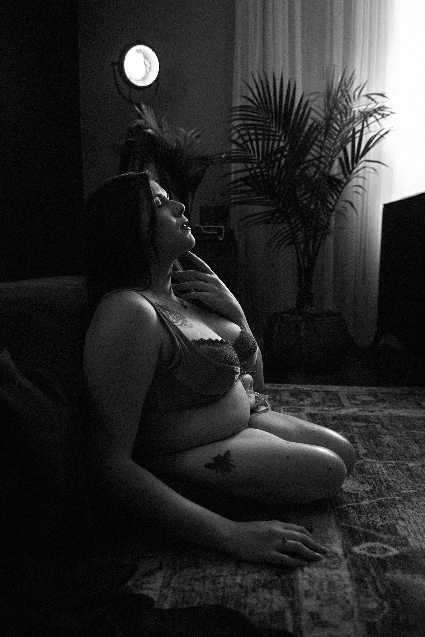 A woman with tattoos on her arm and thigh, wearing lingerie, sitting on the floor in a dimly lit room with a curtain, a potted plant, and a standing lamp in the background.