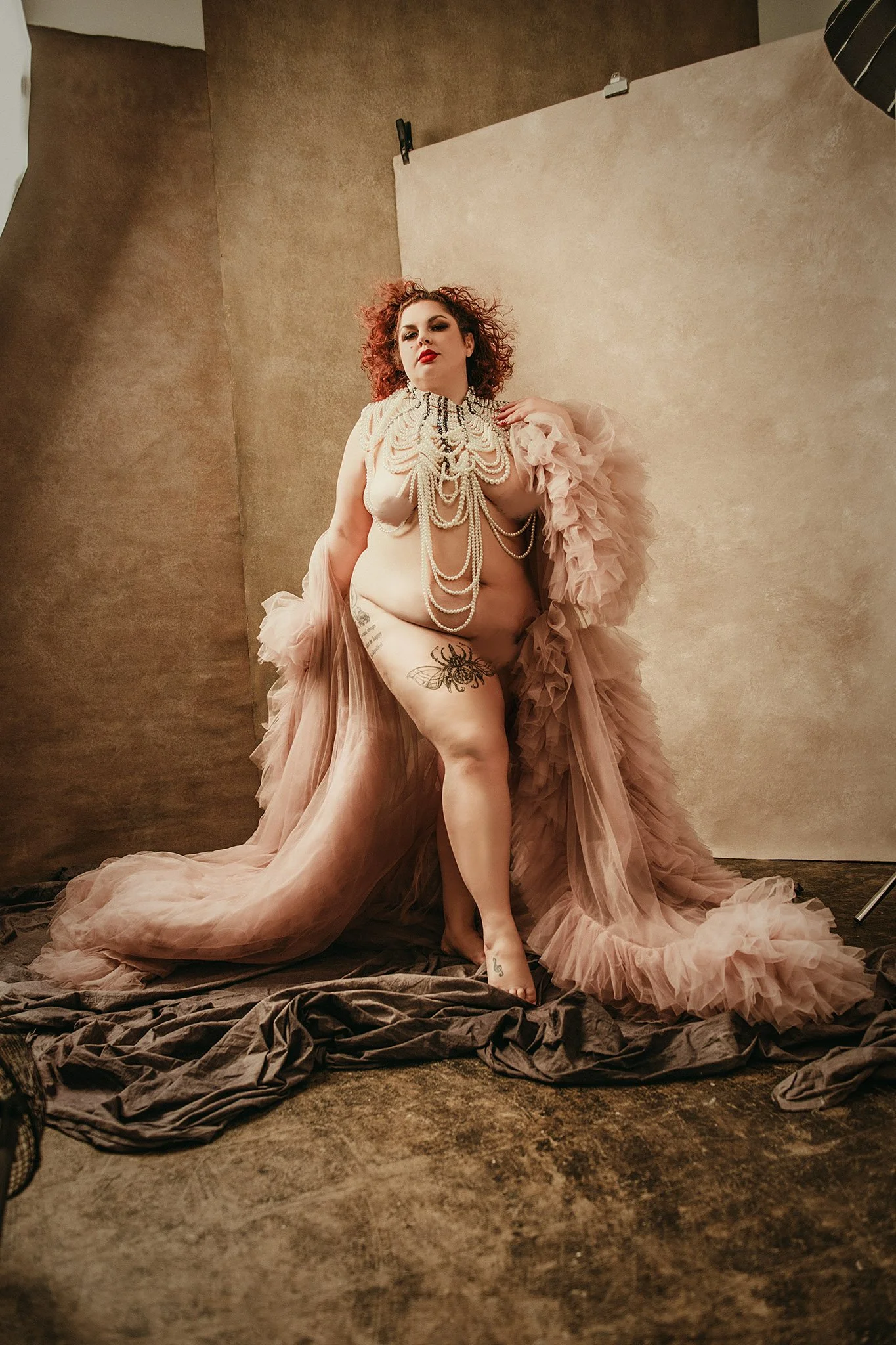 A woman with curly red hair poses in a photography studio, wearing only a large statement pearl necklace and a flowing beige tulle dress, with tattoos on her legs, against a beige backdrop and brown floor.