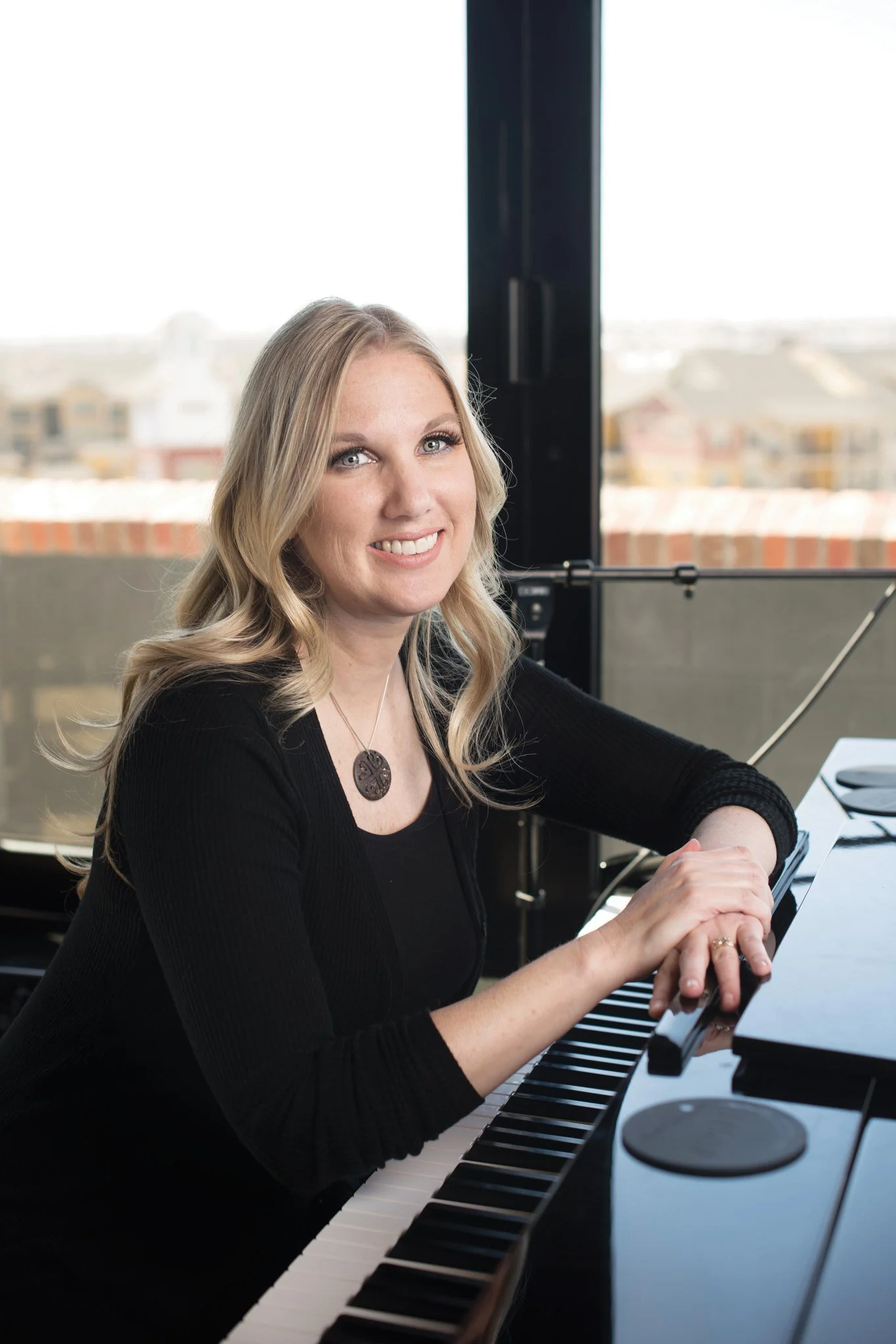 A woman with blonde hair smiling while playing a grand piano in front of large windows with a cityscape view.