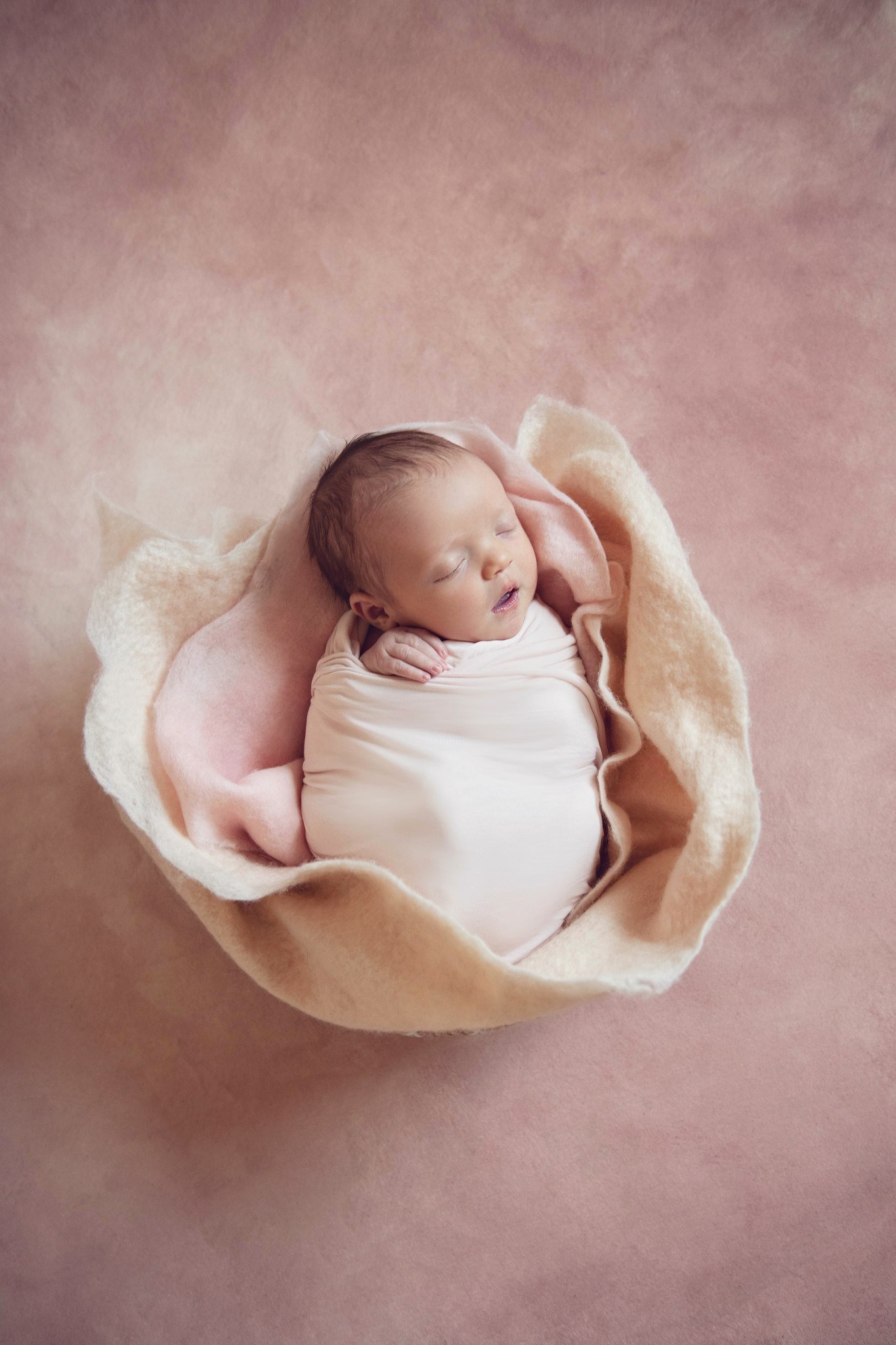 A sleeping newborn baby wrapped snugly in a soft pink blanket, placed in a beige bowl on a pink background.