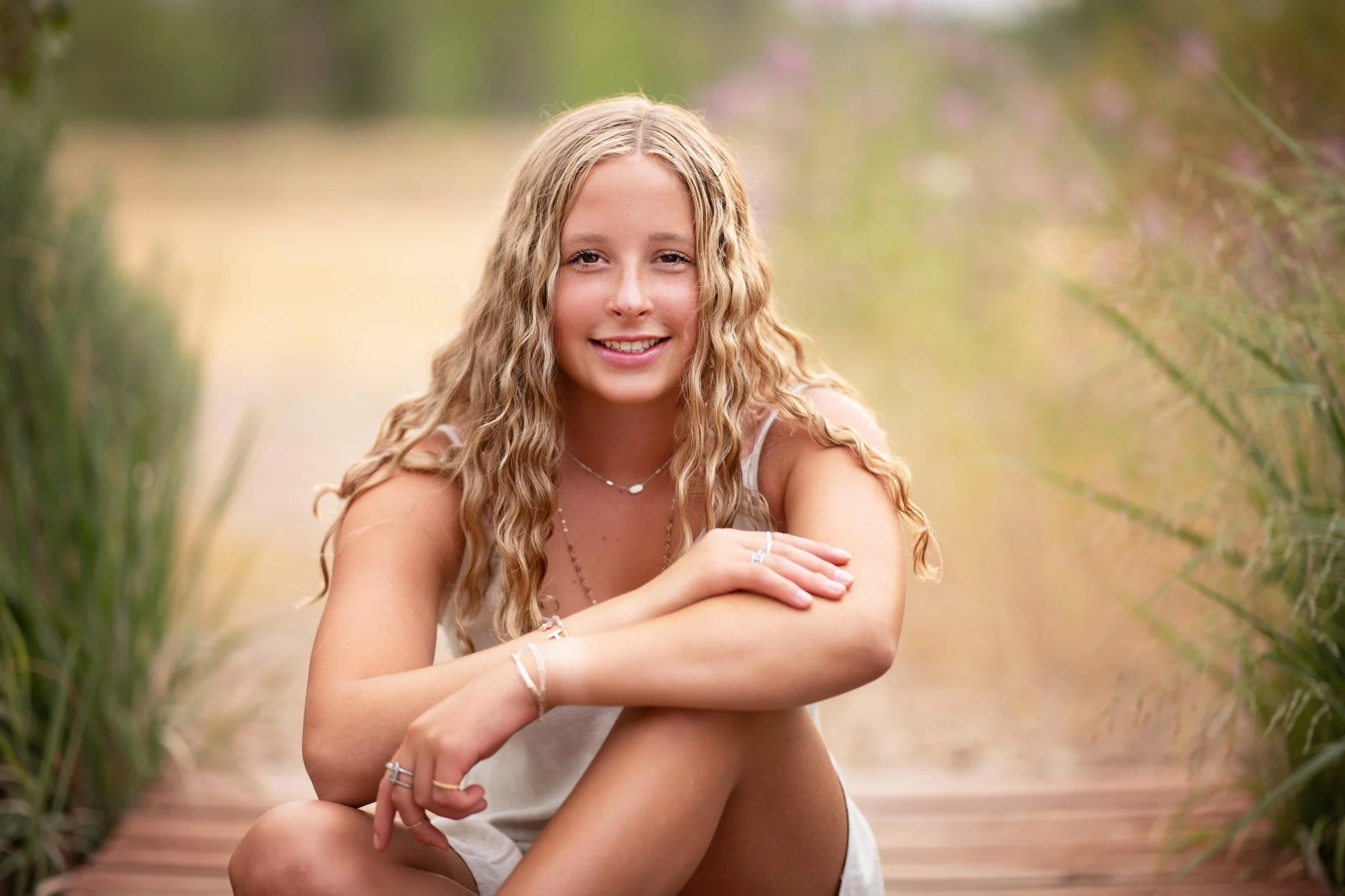 A young woman with long, wavy blonde hair smiling and sitting outdoors on a wooden pathway surrounded by greenery and pink flowers.